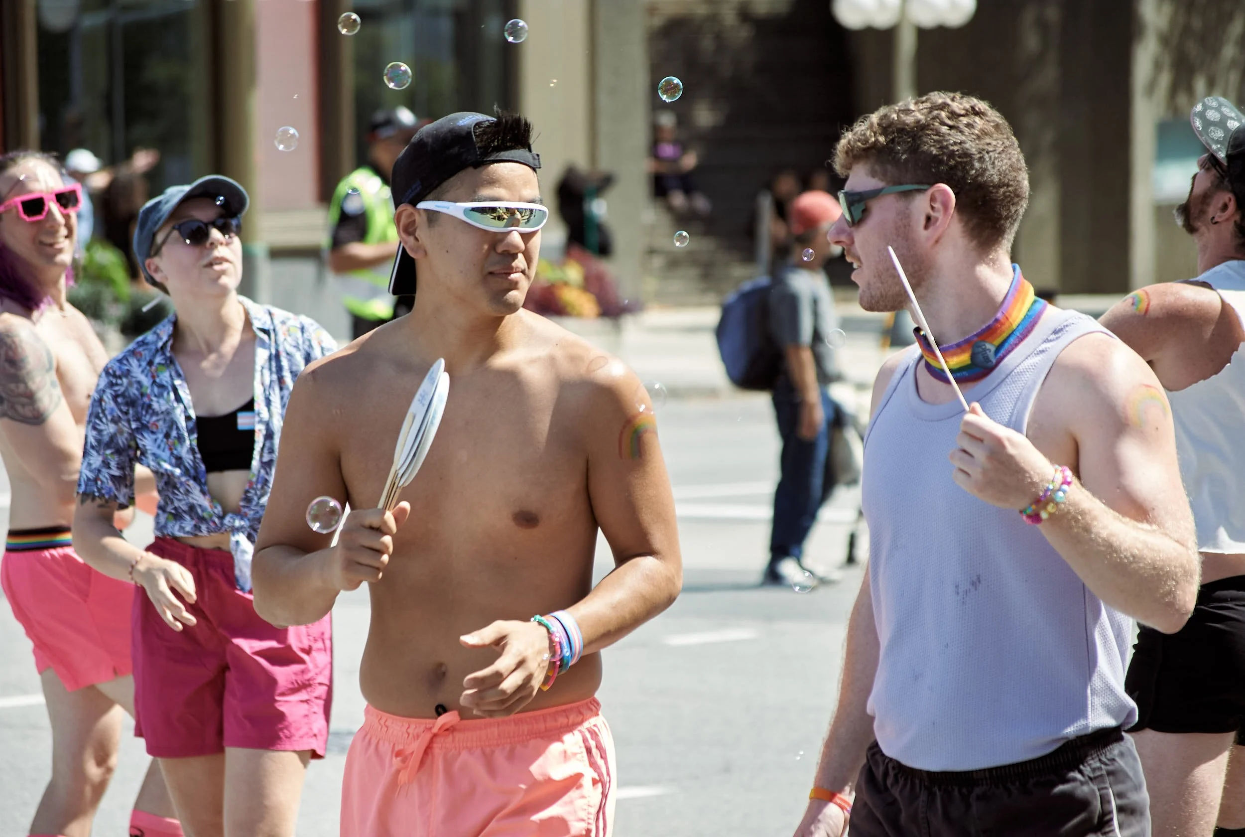 Group of people at pride parade, two men in foreground wearing sunglasses, one shirtless with rainbow wristbands, one in white tank top with rainbow necklace, talking. Others in background wearing colorful clothing and sunglasses, some with rainbow a