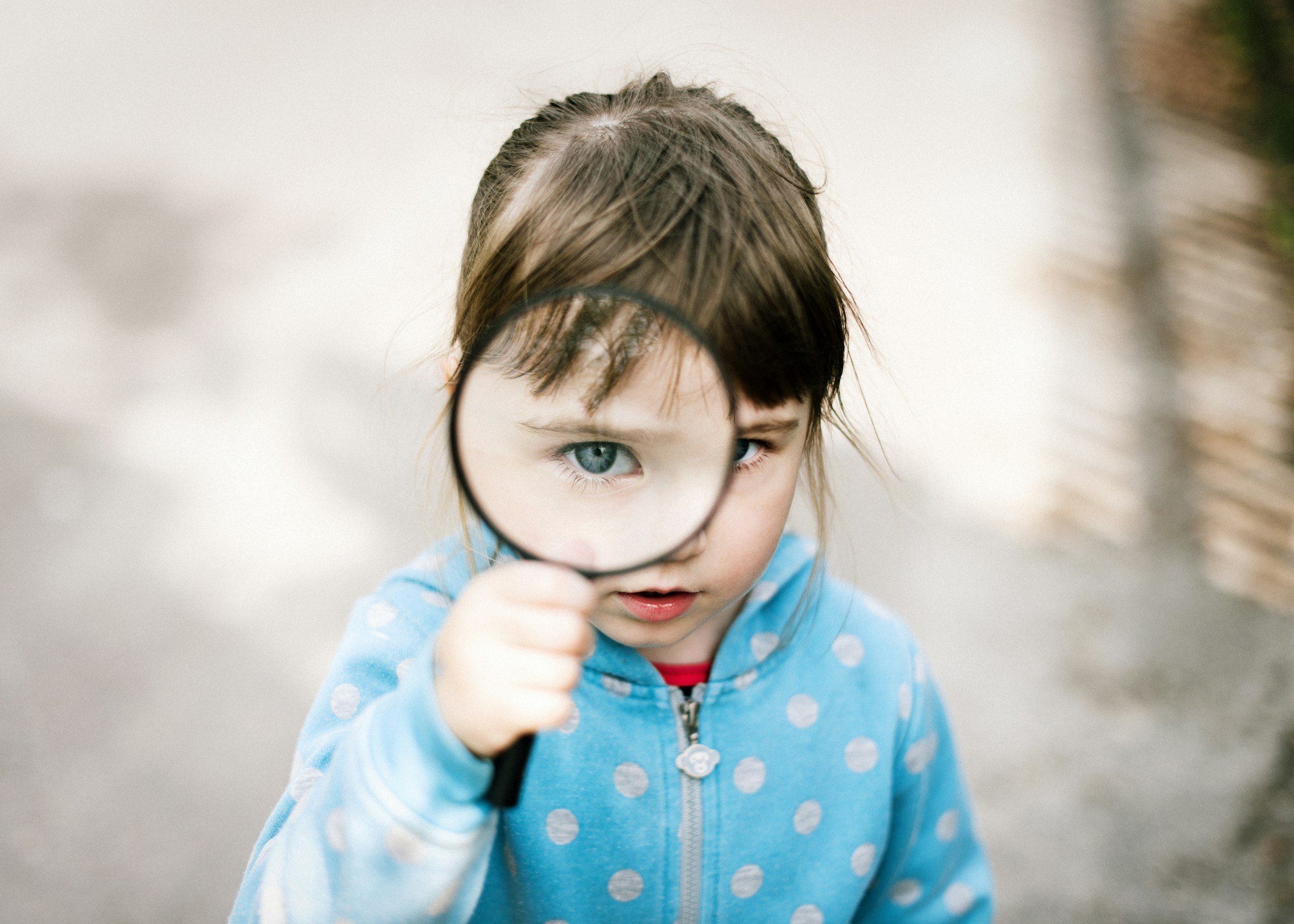 A young girl in a blue polka dot hoodie holds a magnifying glass, examining her eye closely while outdoors.