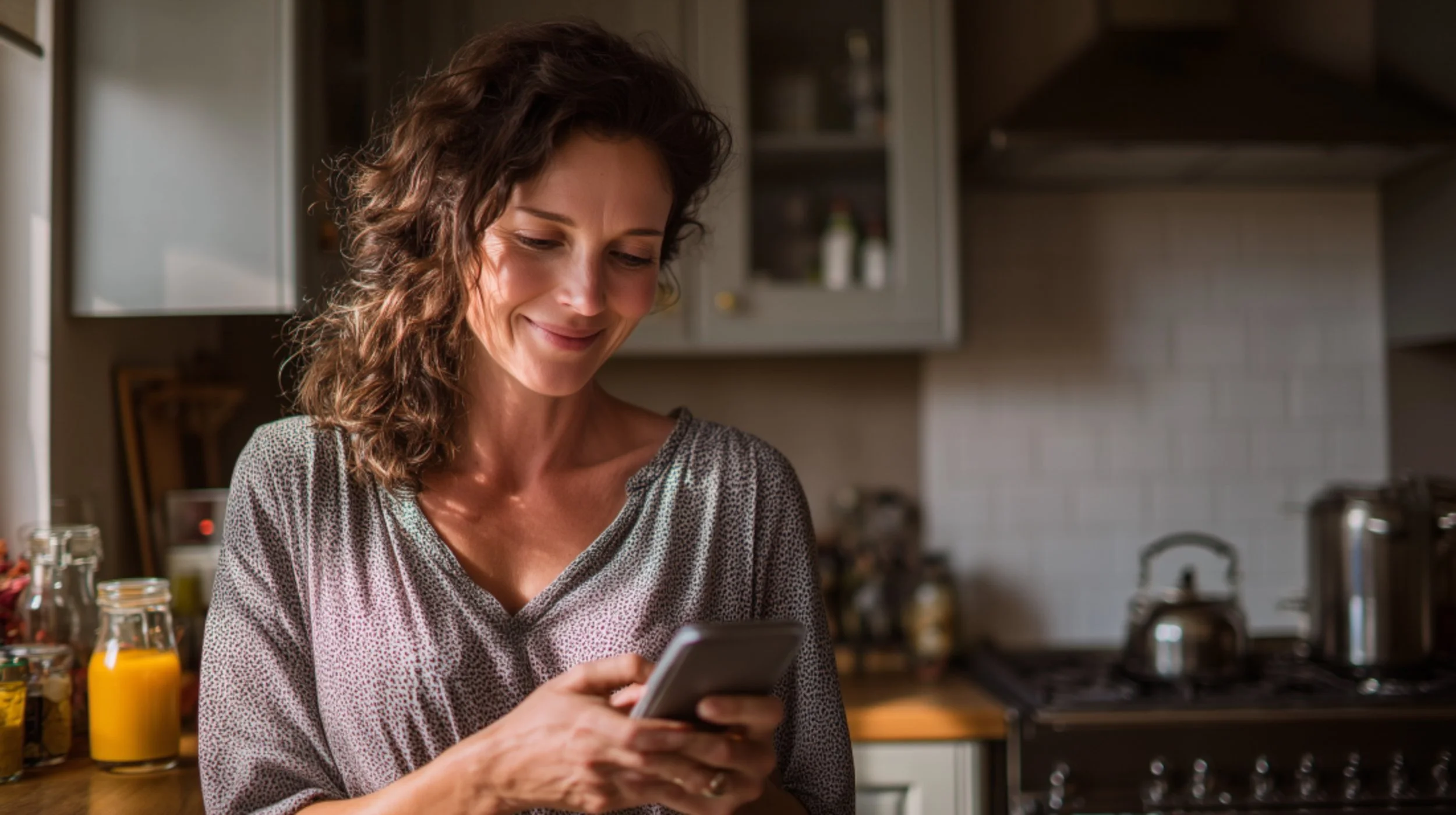 A woman with curly brown hair smiling at her phone in a kitchen with yellow jars and stainless steel appliances in the background.