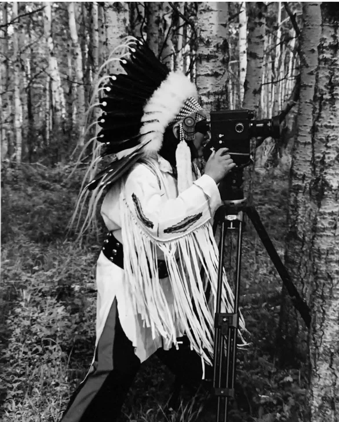 A person dressed in Native American-inspired clothing using a vintage film camera on a tripod in a forest.