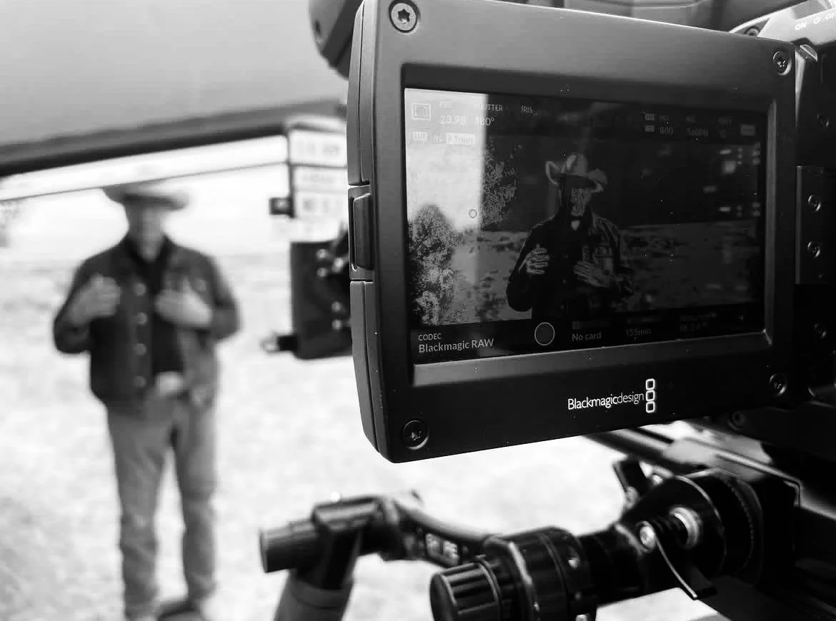 A person dressed as a cowboy is standing outdoors, seen through a camera display, which shows a black-and-white image of the cowboy signifying a photo being taken.