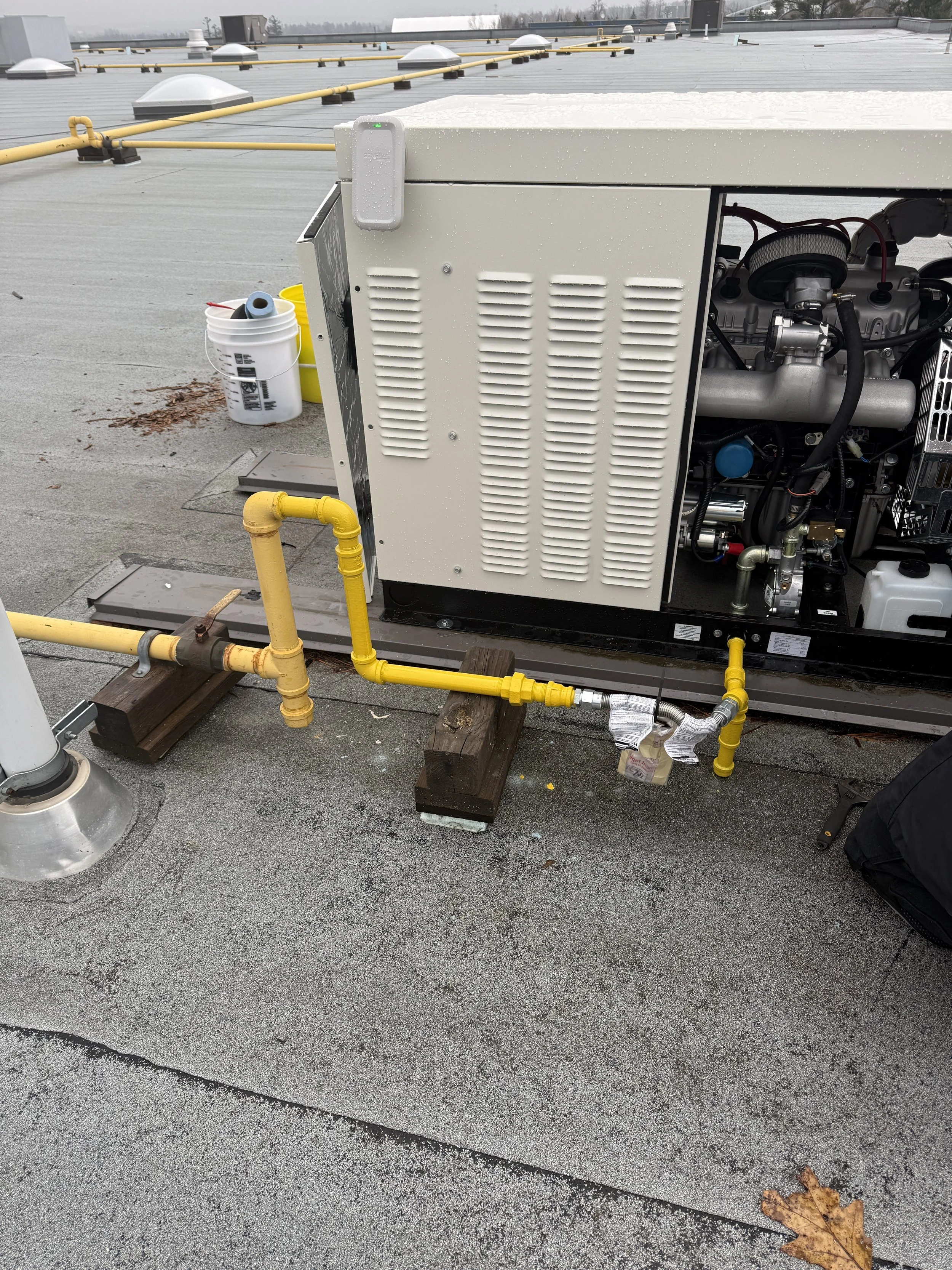 Rooftop HVAC unit with yellow gas pipeline and tools nearby, on a flat roof with some leaves and buckets in the background.