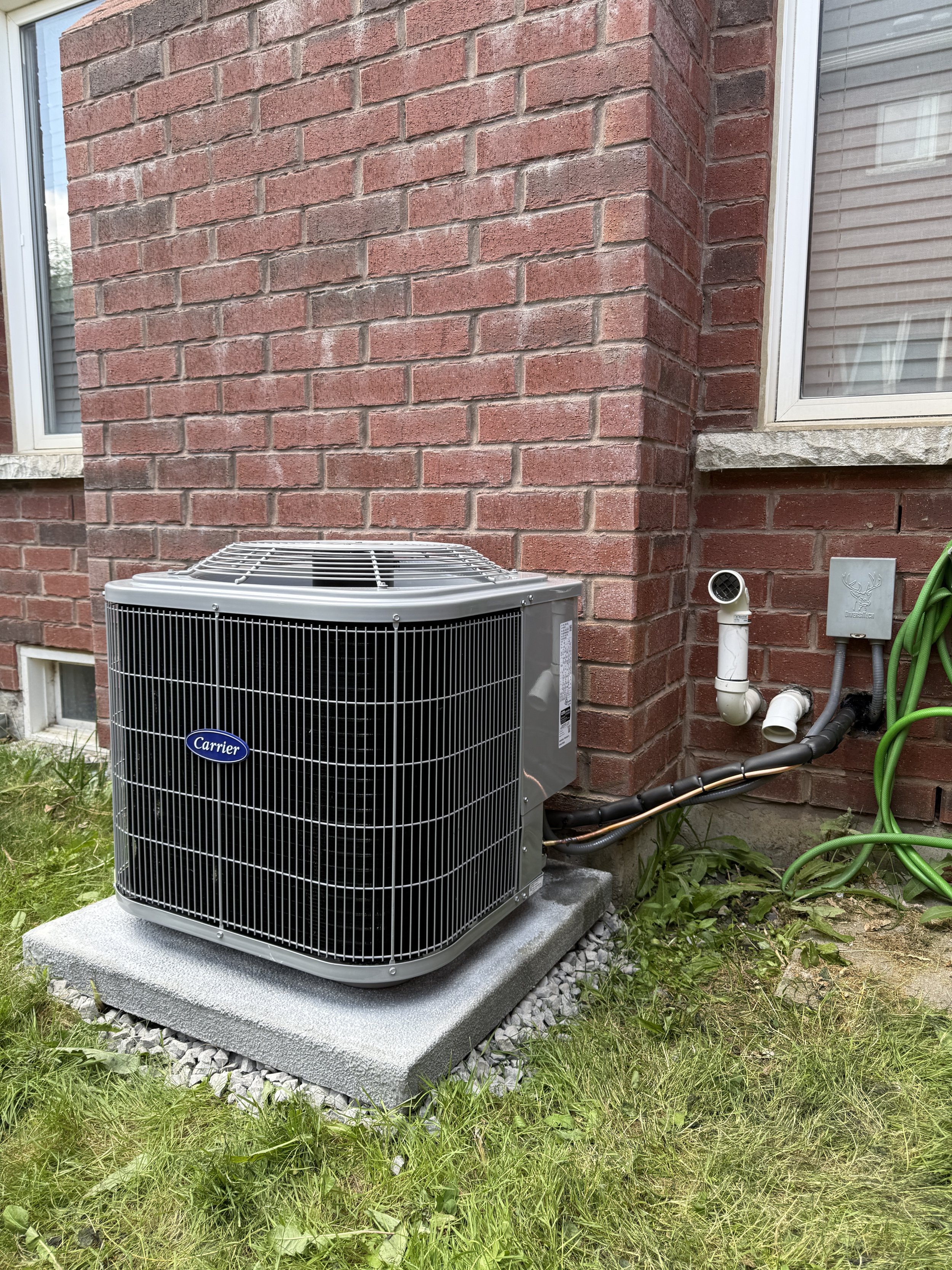 An outdoor air conditioning unit on a concrete pad next to a brick house wall, with green garden hose coiled nearby.