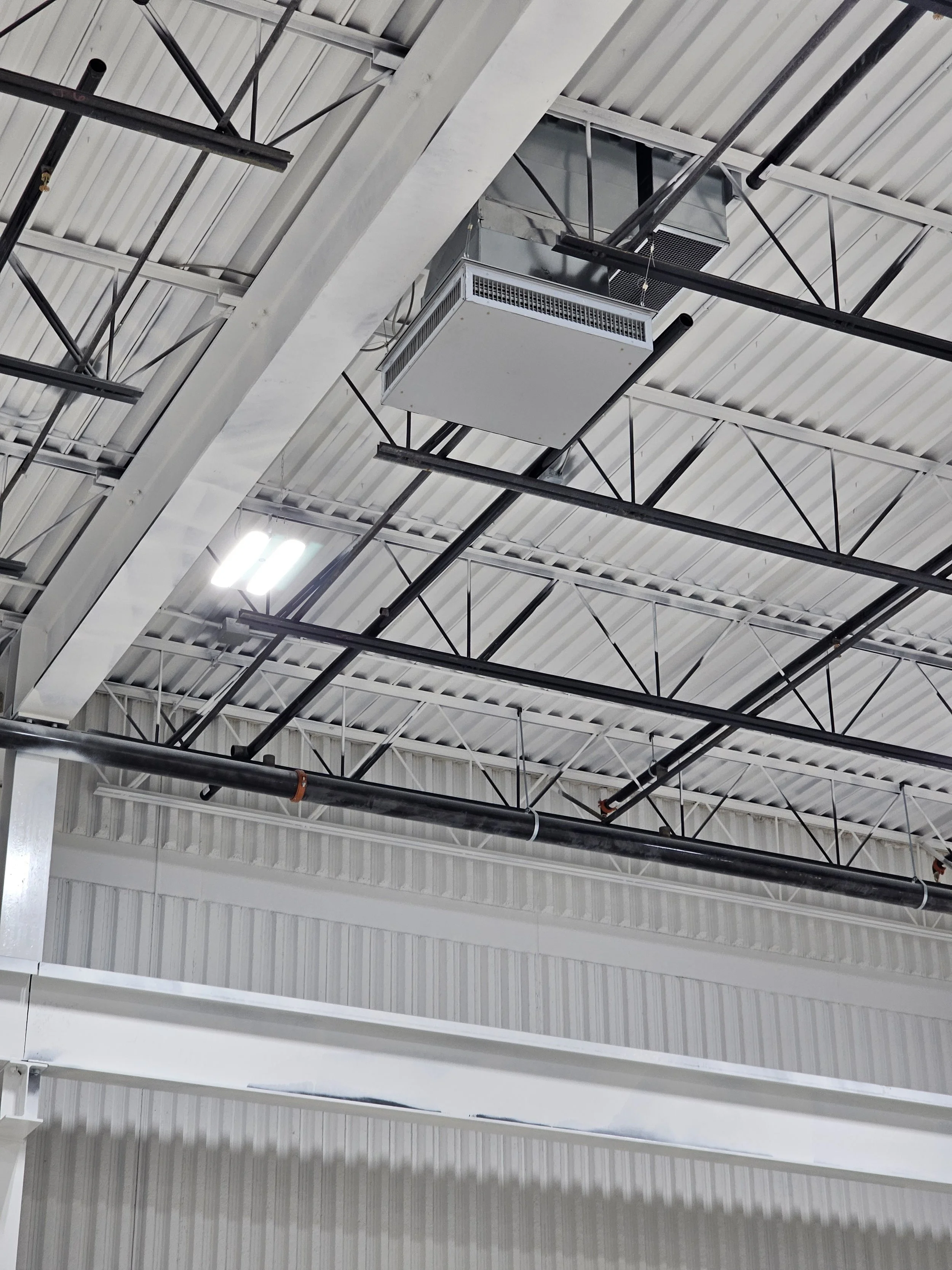 Ceiling with metal beams, a large white air conditioning unit, black pipes, and a bright light fixture.
