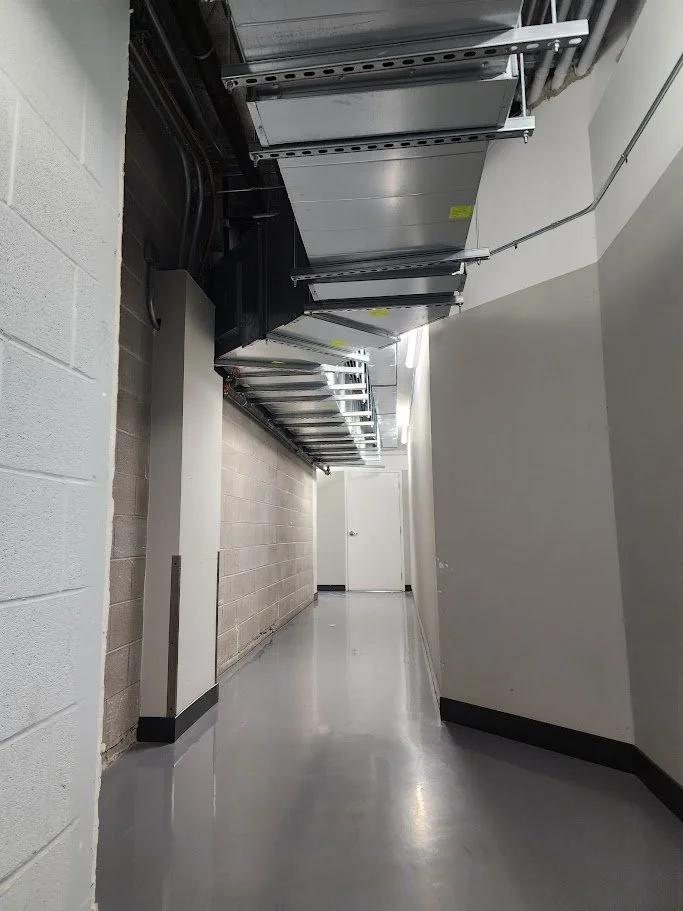 Empty hallway with a gray concrete wall on the left and plain white wall on the right, shiny gray floor, ceiling with metal ductwork and an overhead ladder, white door at the end of the hallway.
