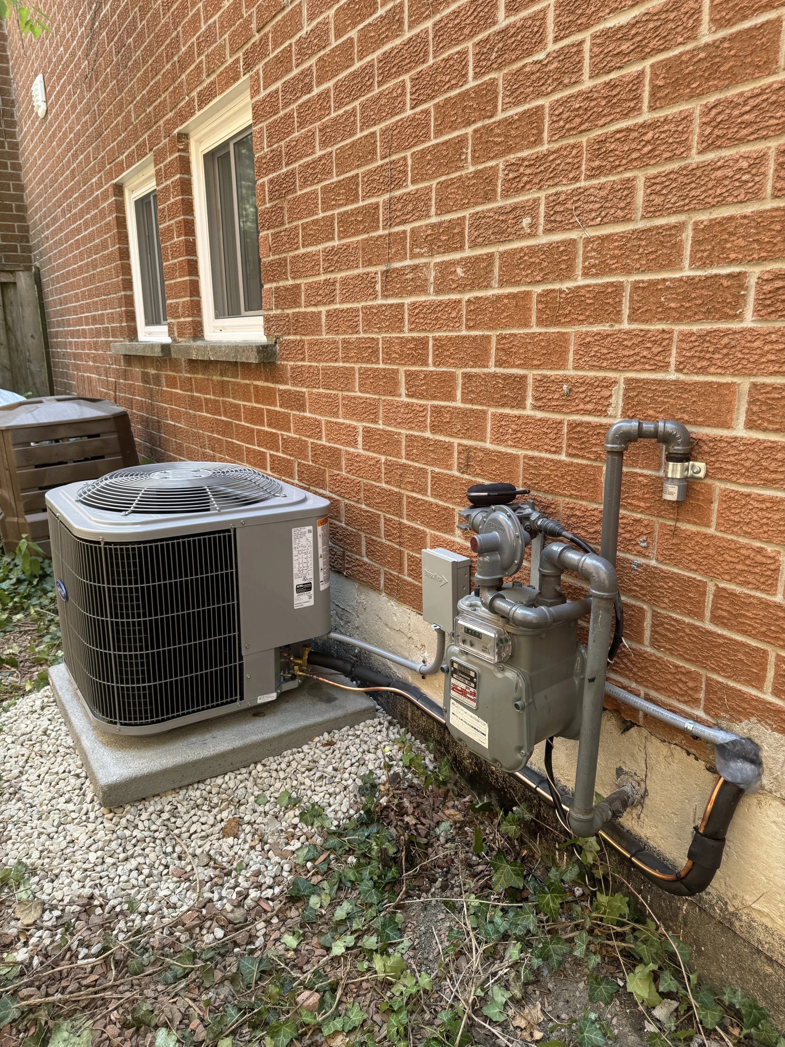 The image shows the outdoor air conditioning unit and gas meter installed outside a brick house wall. The air conditioning unit is on a concrete slab on the ground, with a gravel and ivy-covered area surrounding it. The gas meter and piping are mount