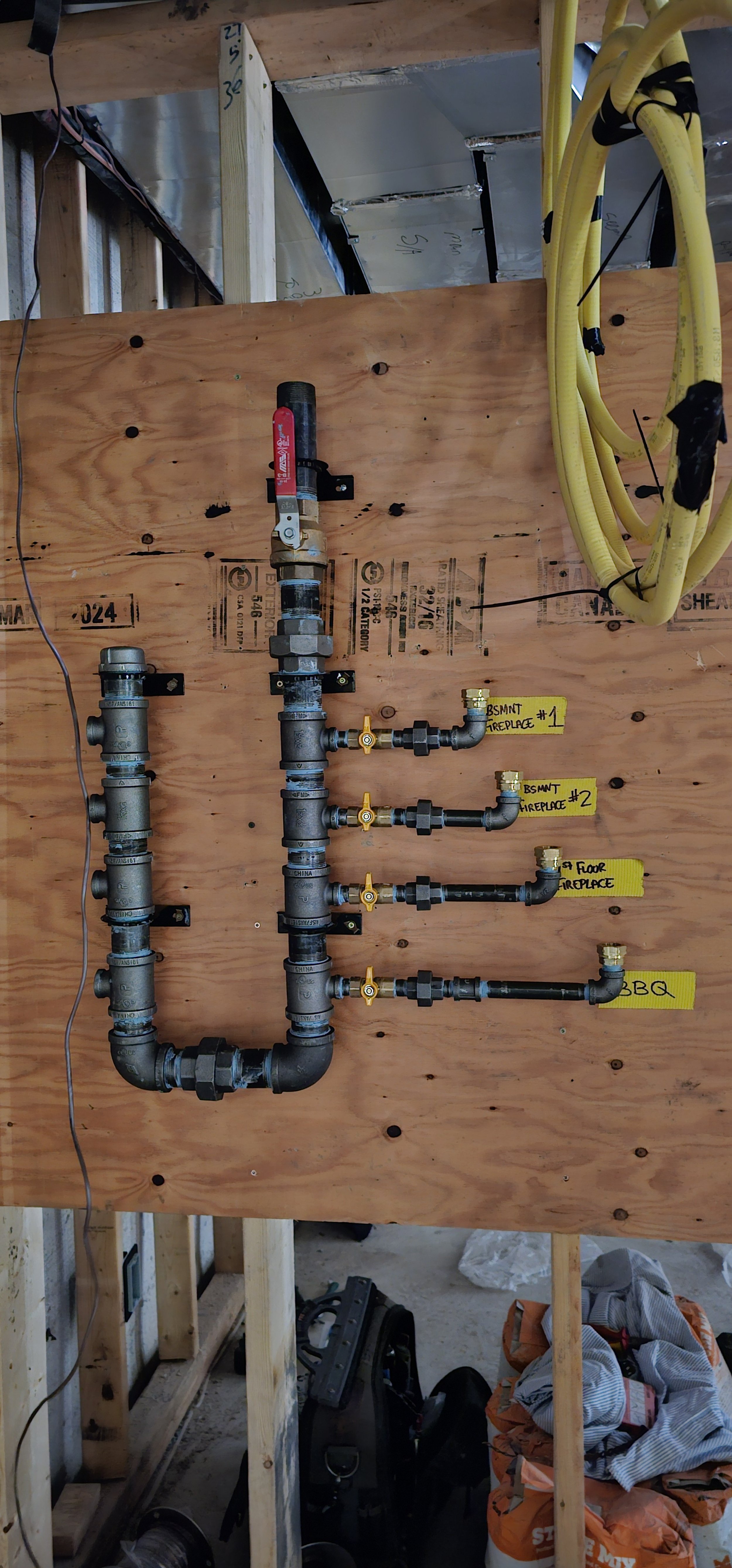 Water pipe fittings on a wooden panel labeled with yellow tags for different heating zones, including fireplace and BBQ.