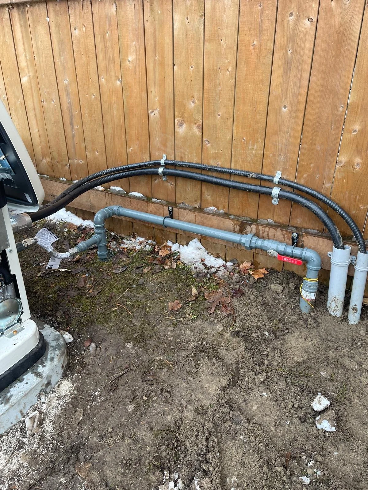 A section of a house foundation with exposed utility pipes, installed along a wooden fence, with dirt and some lingering snow on the ground.