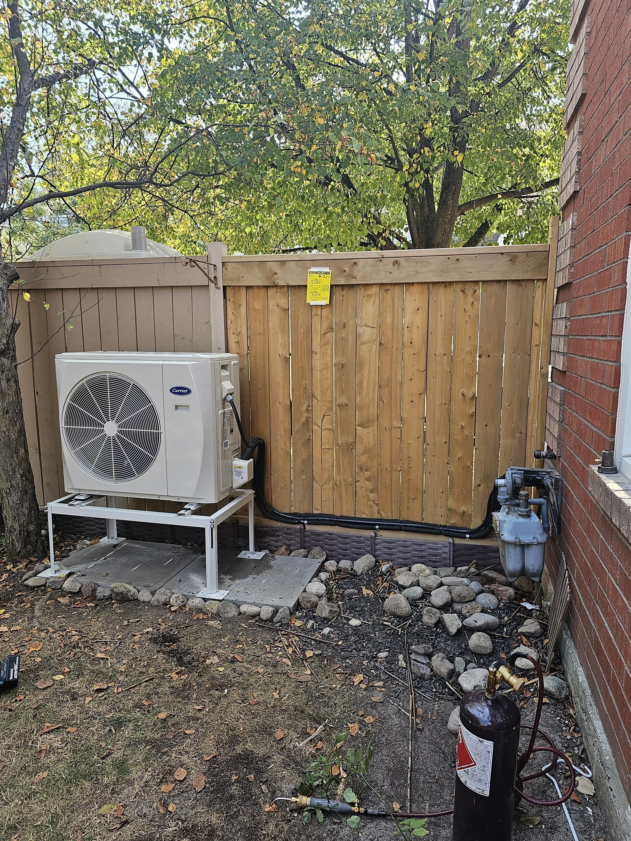 A backyard scene with an HVAC unit installed on a small concrete pad in front of a wooden privacy fence, next to a red brick house wall, with a tree providing shade and a gas meter nearby.