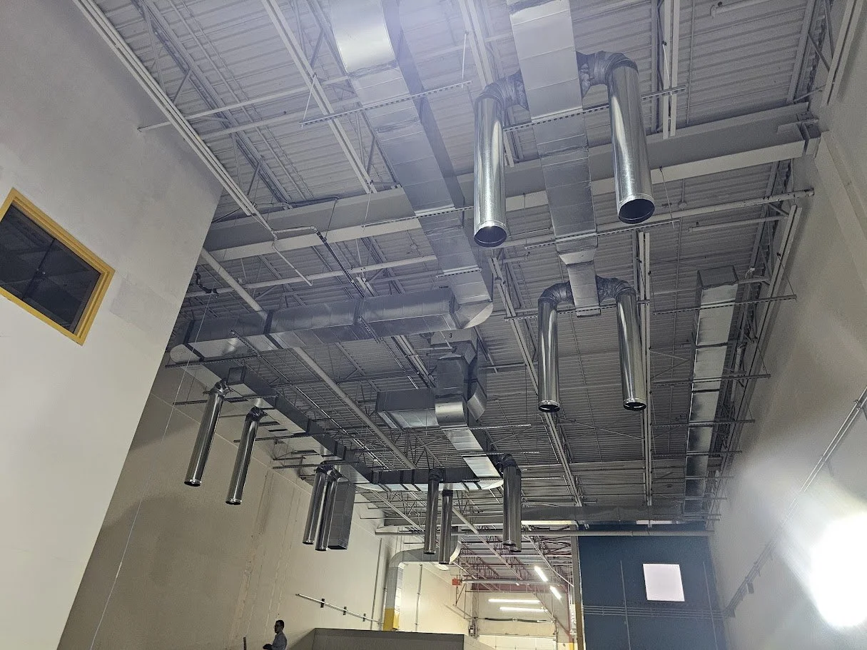 View of indoor ceiling with extensive metal ductwork, ventilation pipes, and exposed ceiling structure in a commercial or industrial space.