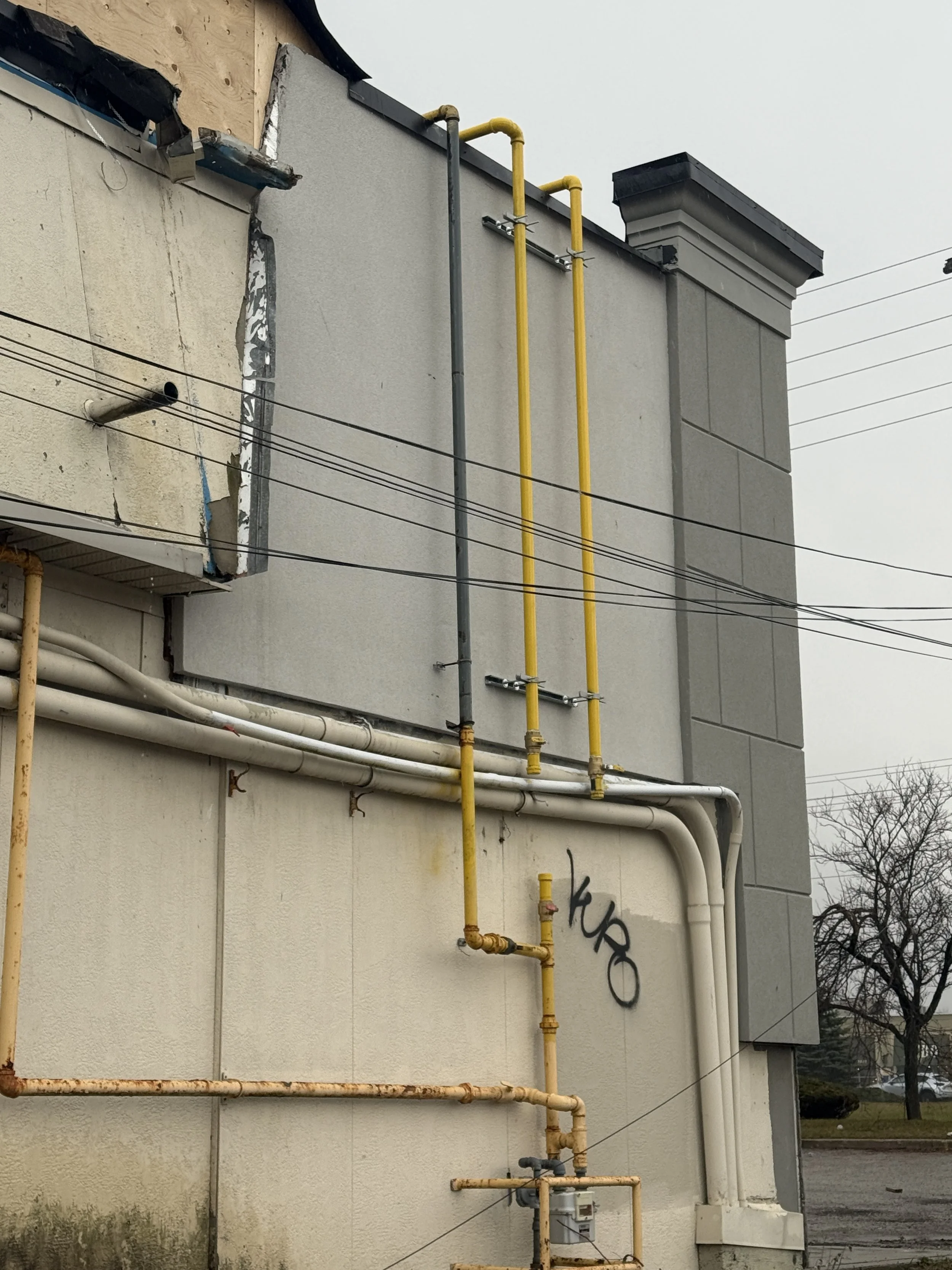 Side of a building with exposed yellow and white pipes, graffiti on the wall, and a gray cloudy sky in the background.