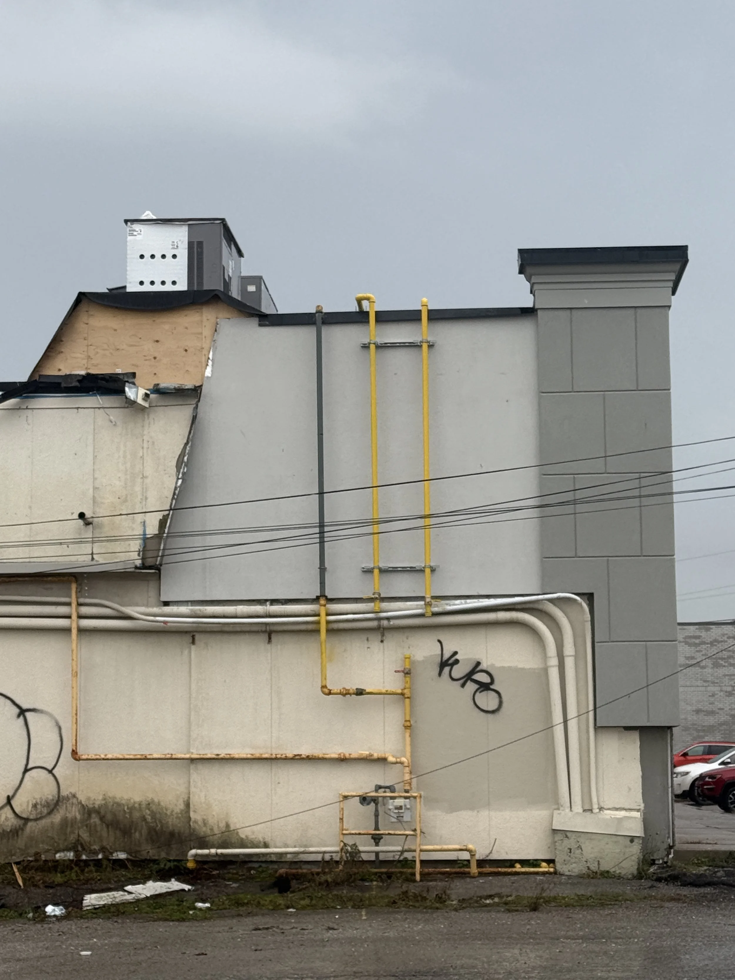 The side of a building with visible yellow and gray gas pipes, graffiti, and a cloudy sky in the background.