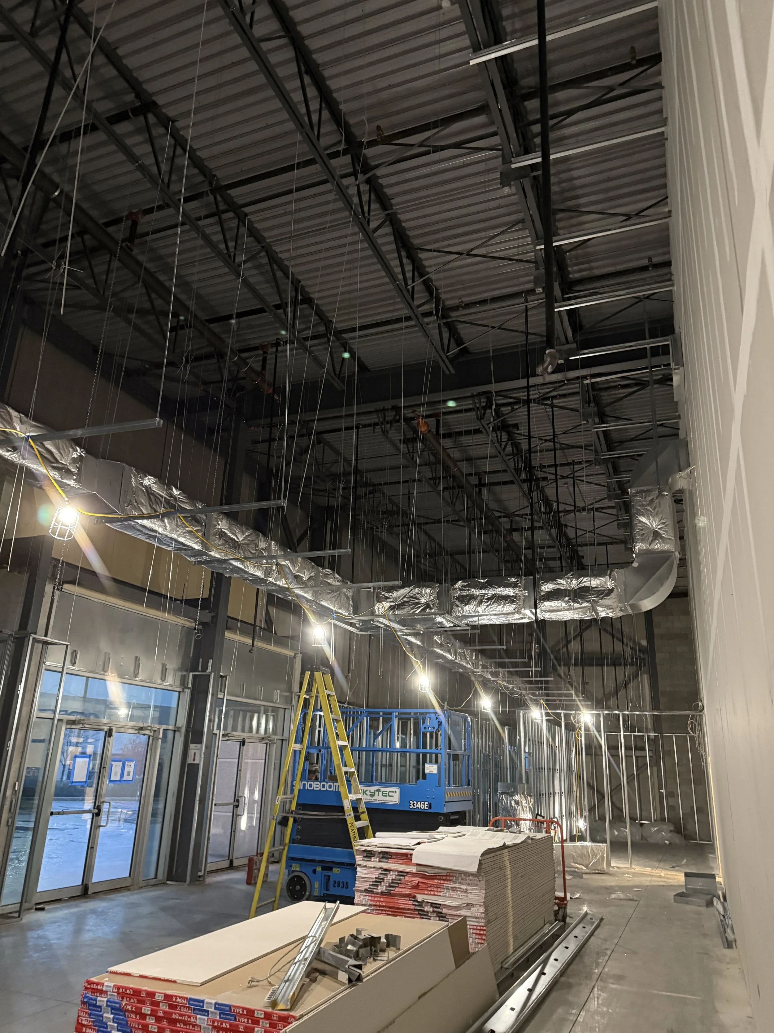 Construction site inside a building with a high ceiling, exposed metal framework, ductwork, and construction equipment including a ladder, a scissor lift, and stacks of materials.