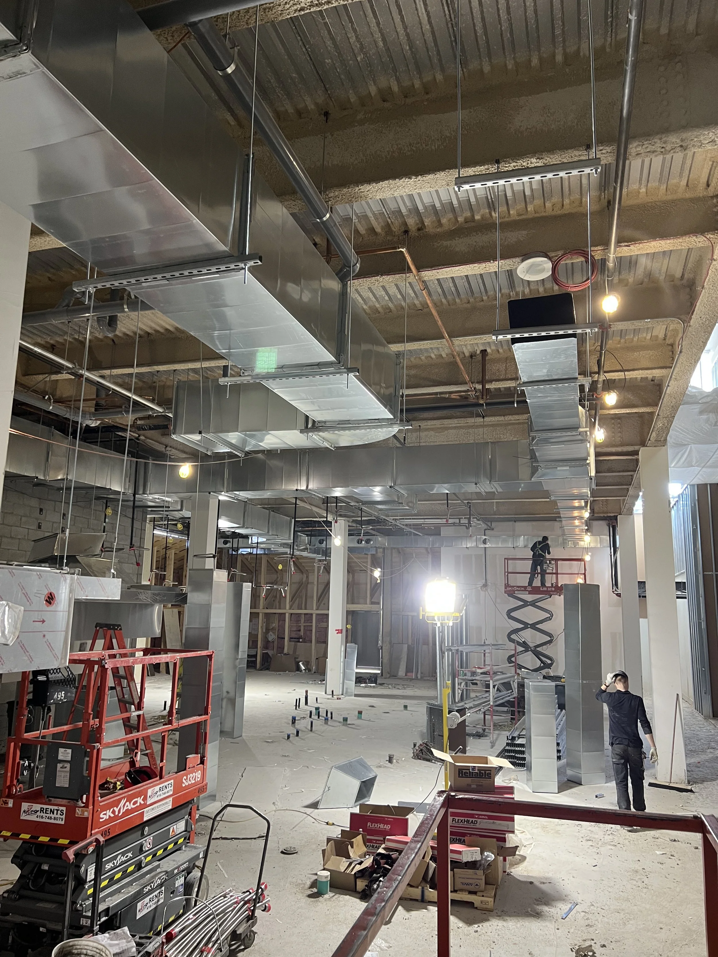 Construction site with metal ductwork installation, workers on lift, and scattered tools and materials.