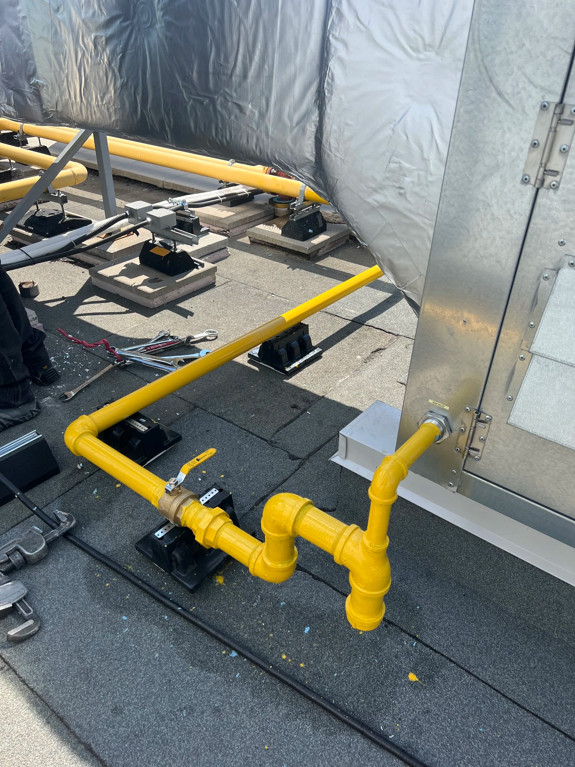 Yellow pipeline with valves on a rooftop, connected to metal equipment and secured with black mounting brackets, with tools and miscellaneous items nearby.