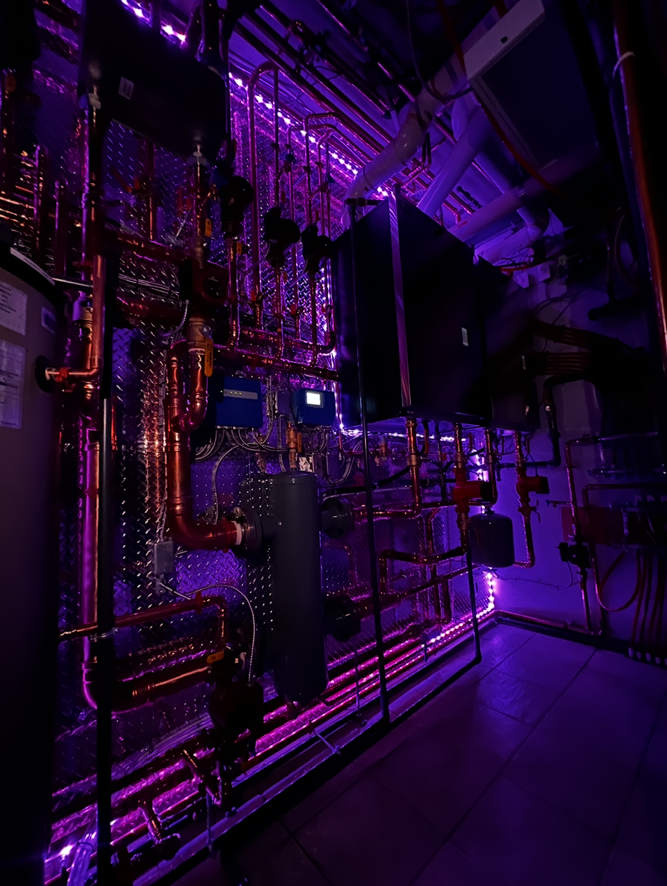 A room with an intricate network of metallic pipes and machinery illuminated by purple LED lights.
