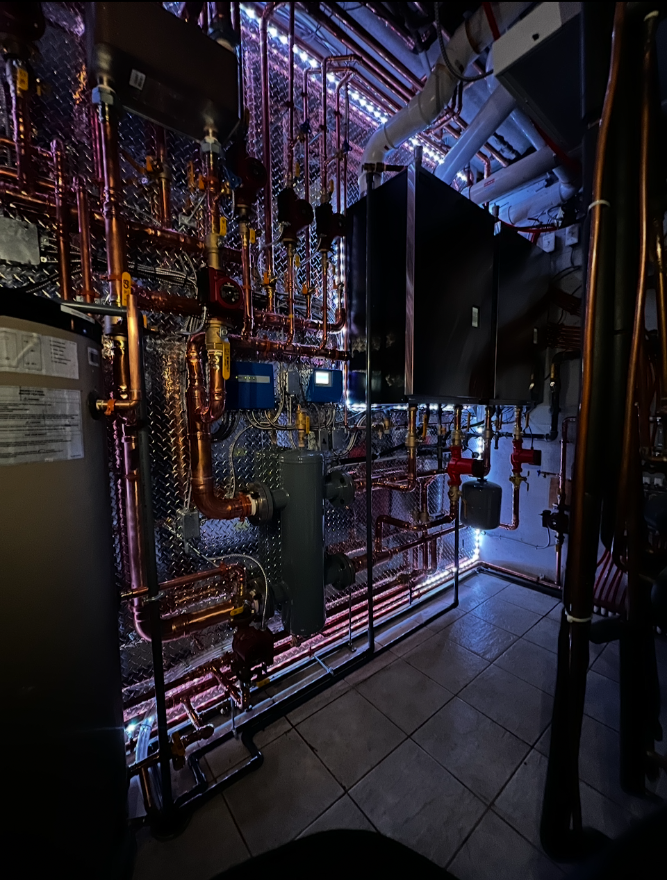 Industrial mechanical room with copper pipes, gauges, and electronic controls illuminated by LED lighting.
