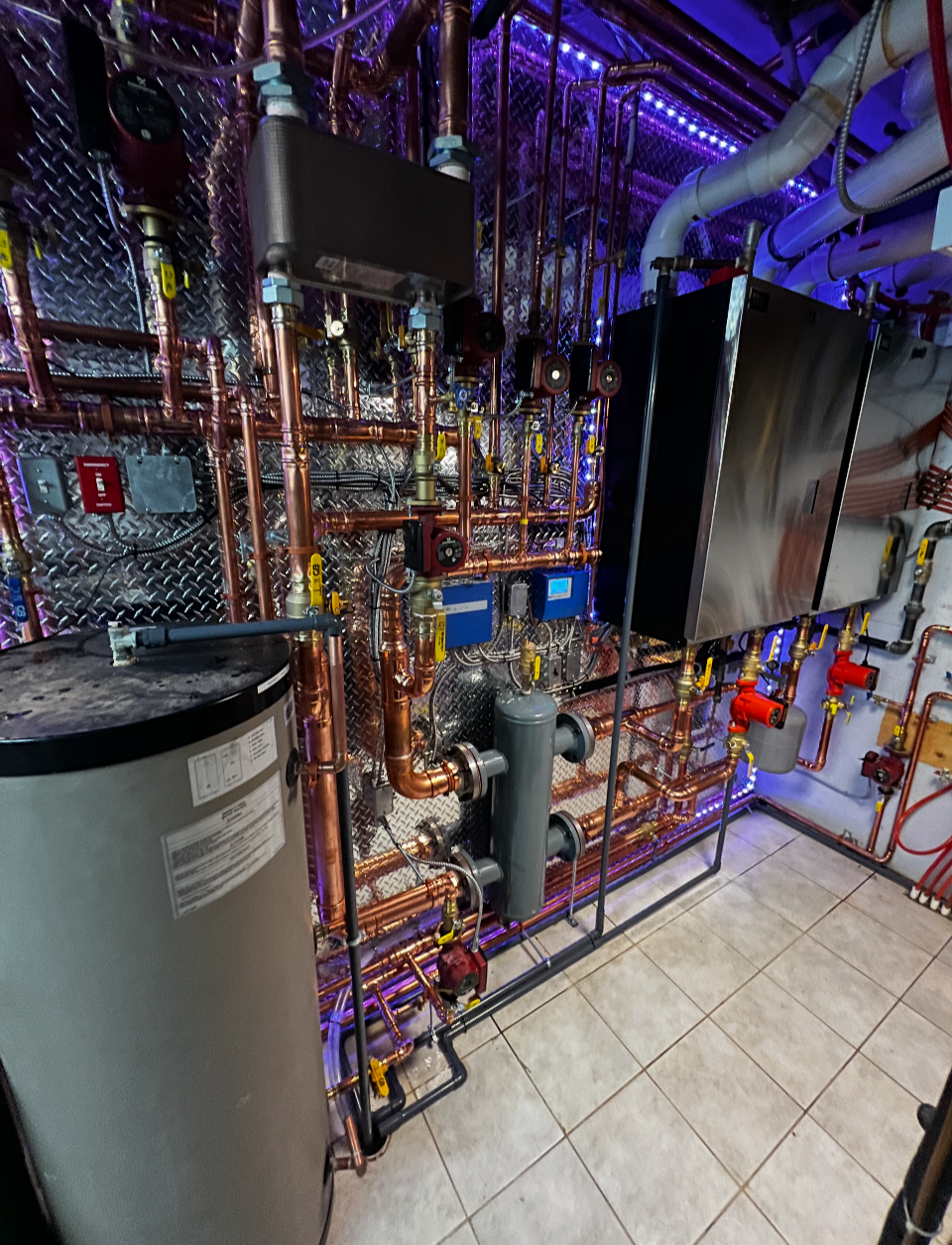 A mechanical room with copper pipes, a large tank, digital controllers, and a black cabinet, illuminated by purple lighting.
