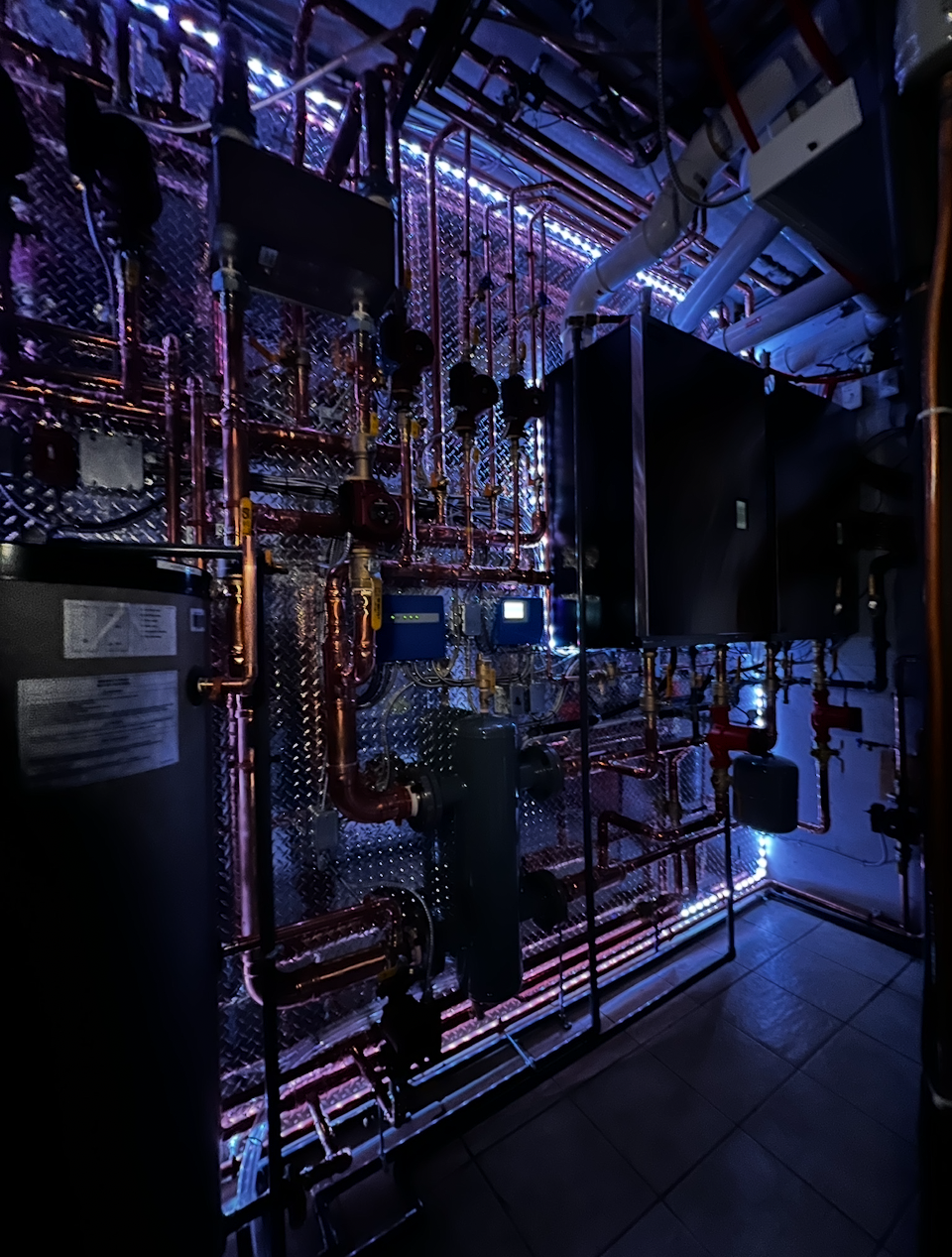 A room with an intricate system of copper pipes, panels, and machinery, illuminated by blue and purple LED lights, likely a mechanical or utility room.