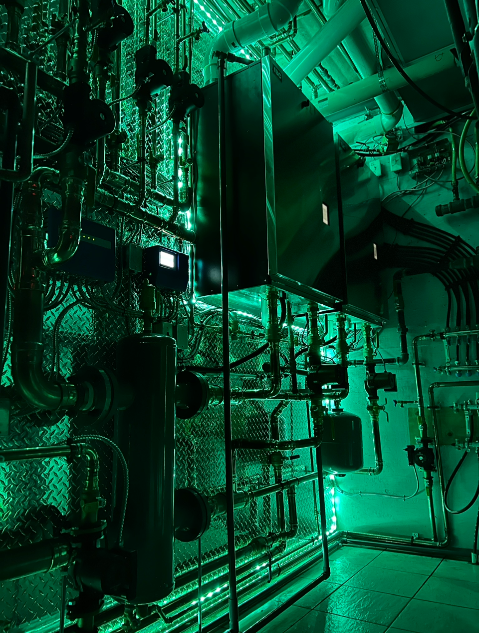 Industrial equipment with black machines and piping, illuminated by green LED lights in a utility room.