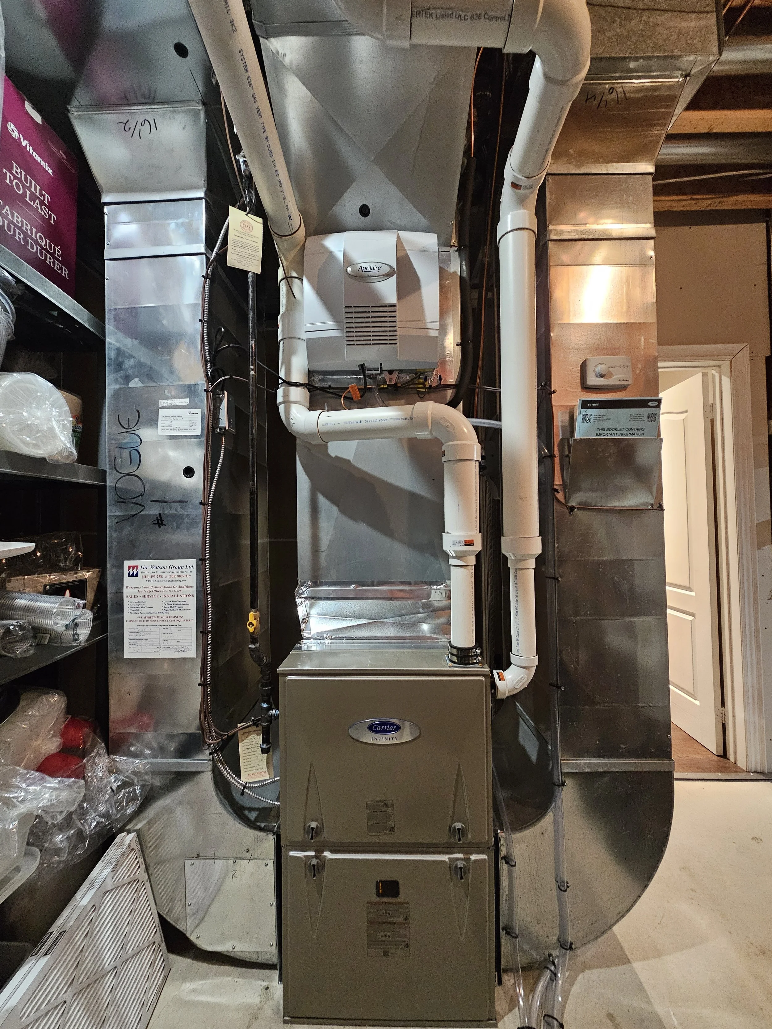 An HVAC furnace system installed in a utility space with metal ducts, PVC pipes, and a control panel, surrounded by shelves with storage items.
