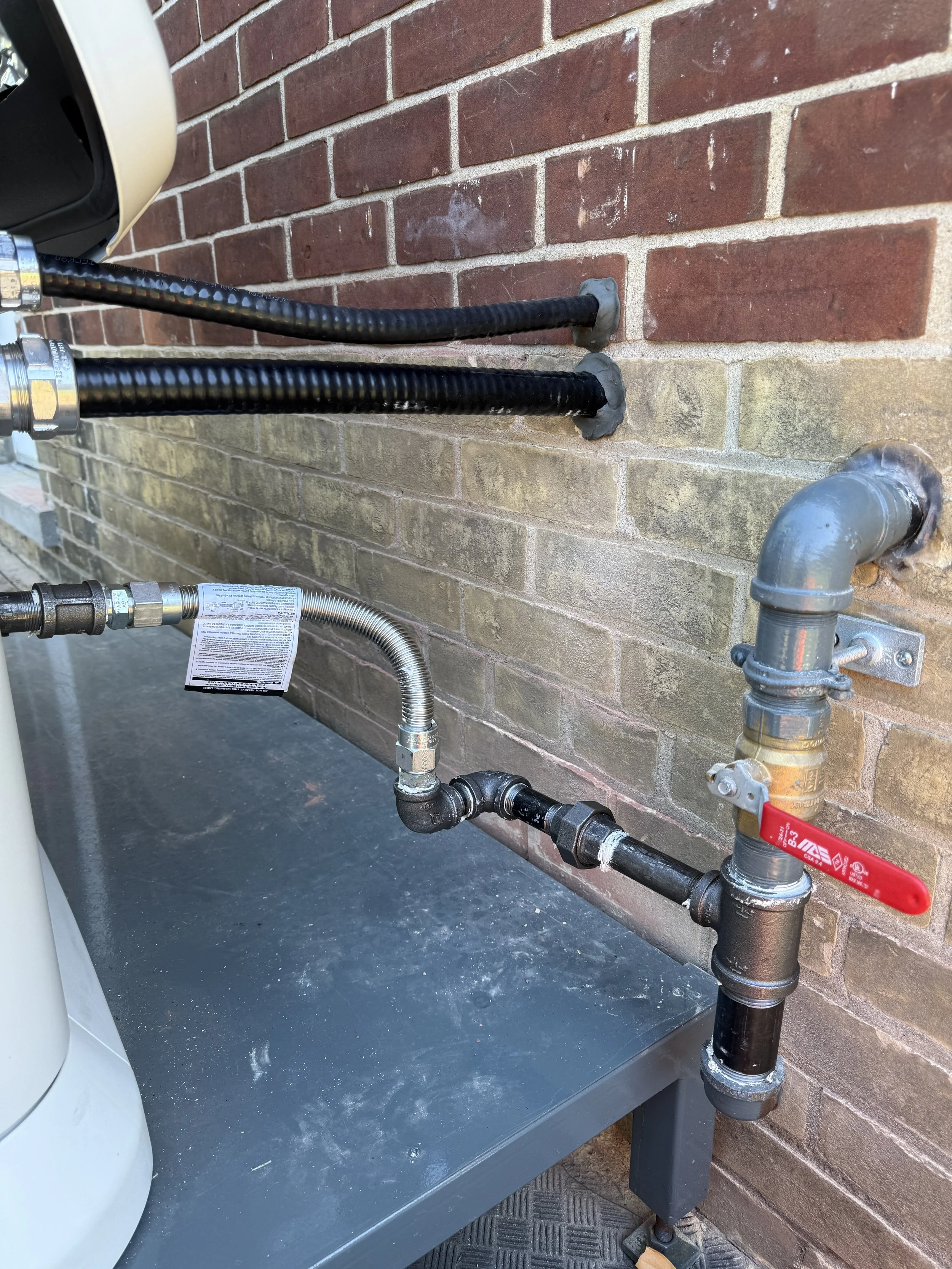 Outdoor plumbing with black and silver pipes, a red valve handle, and a gray table against a brick wall.