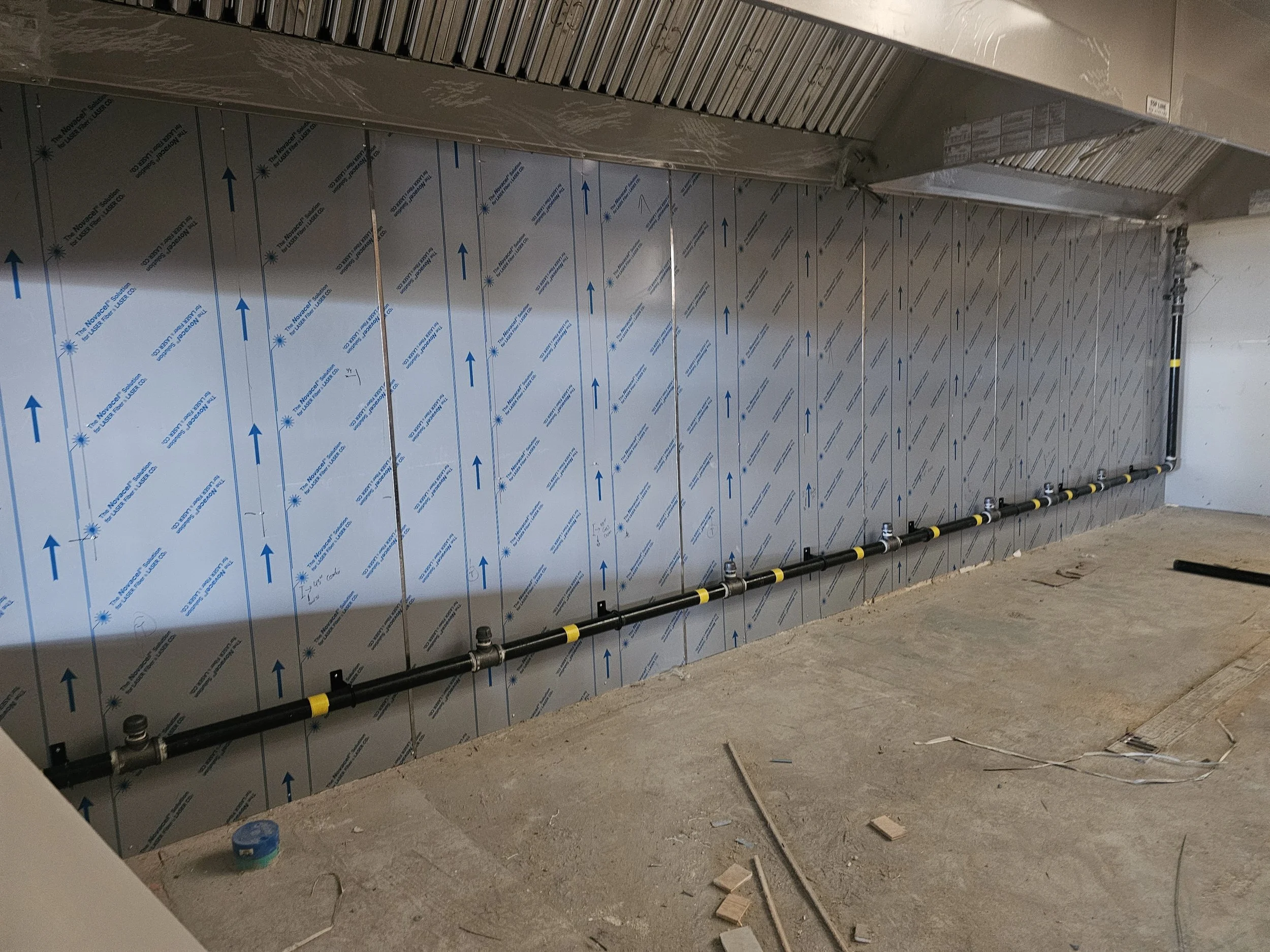 Construction site showing insulated wall with metal ductwork, pipes, and electrical wiring, unfinished floor.