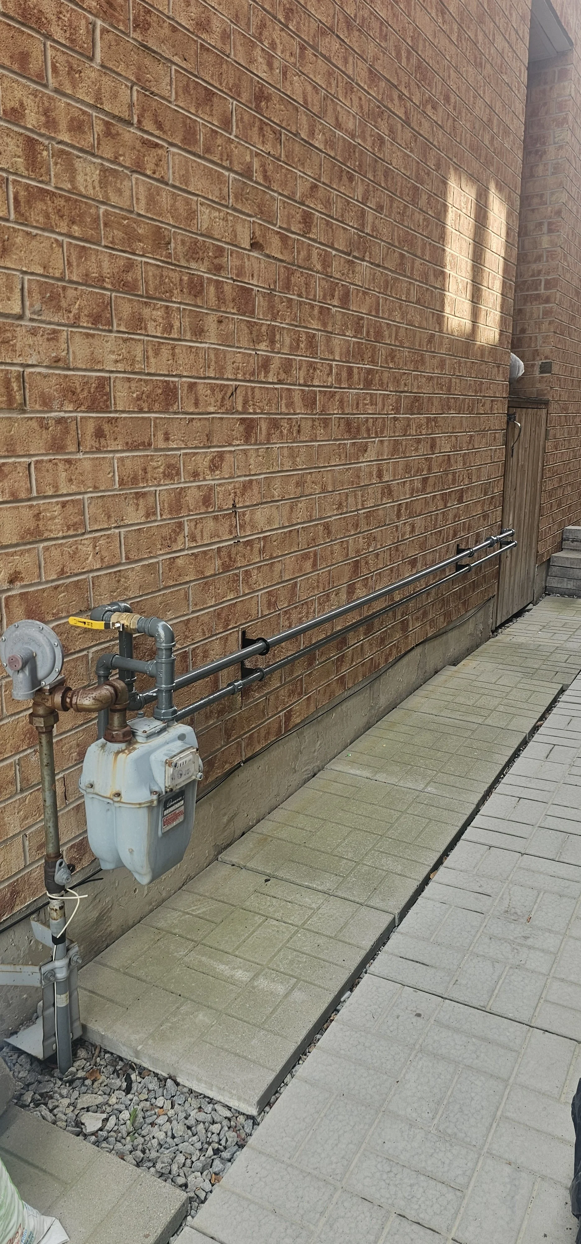 Outdoor brick wall with gas meter and piping, paved pathway with beige and gray tiles.