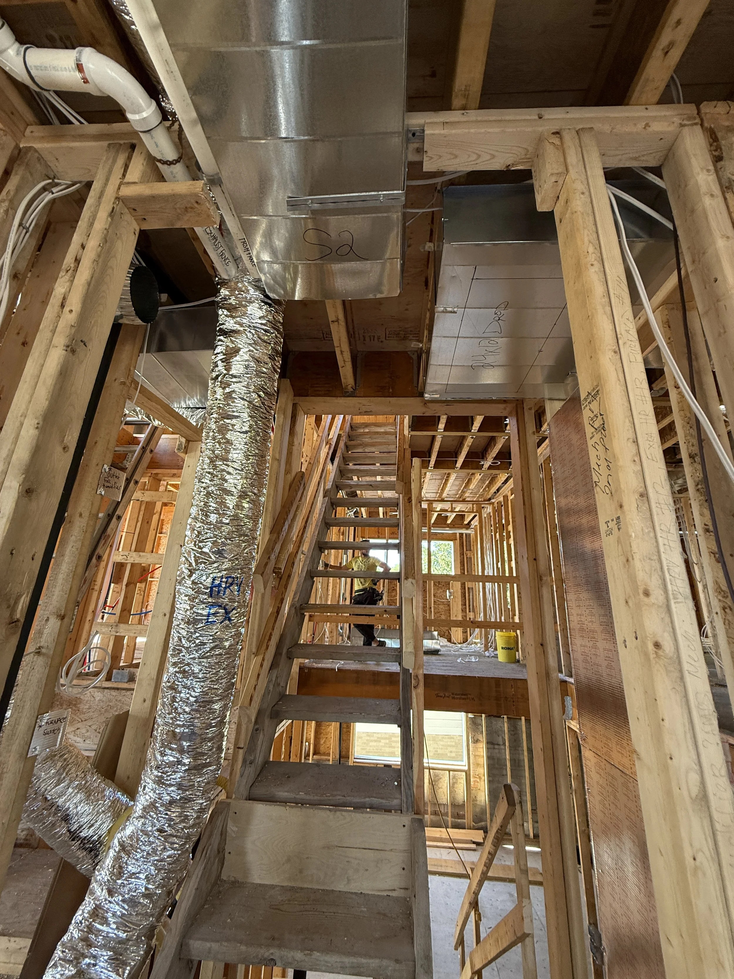 View of the interior of a house under construction, showing exposed wooden framing, a staircase, and HVAC ductwork.