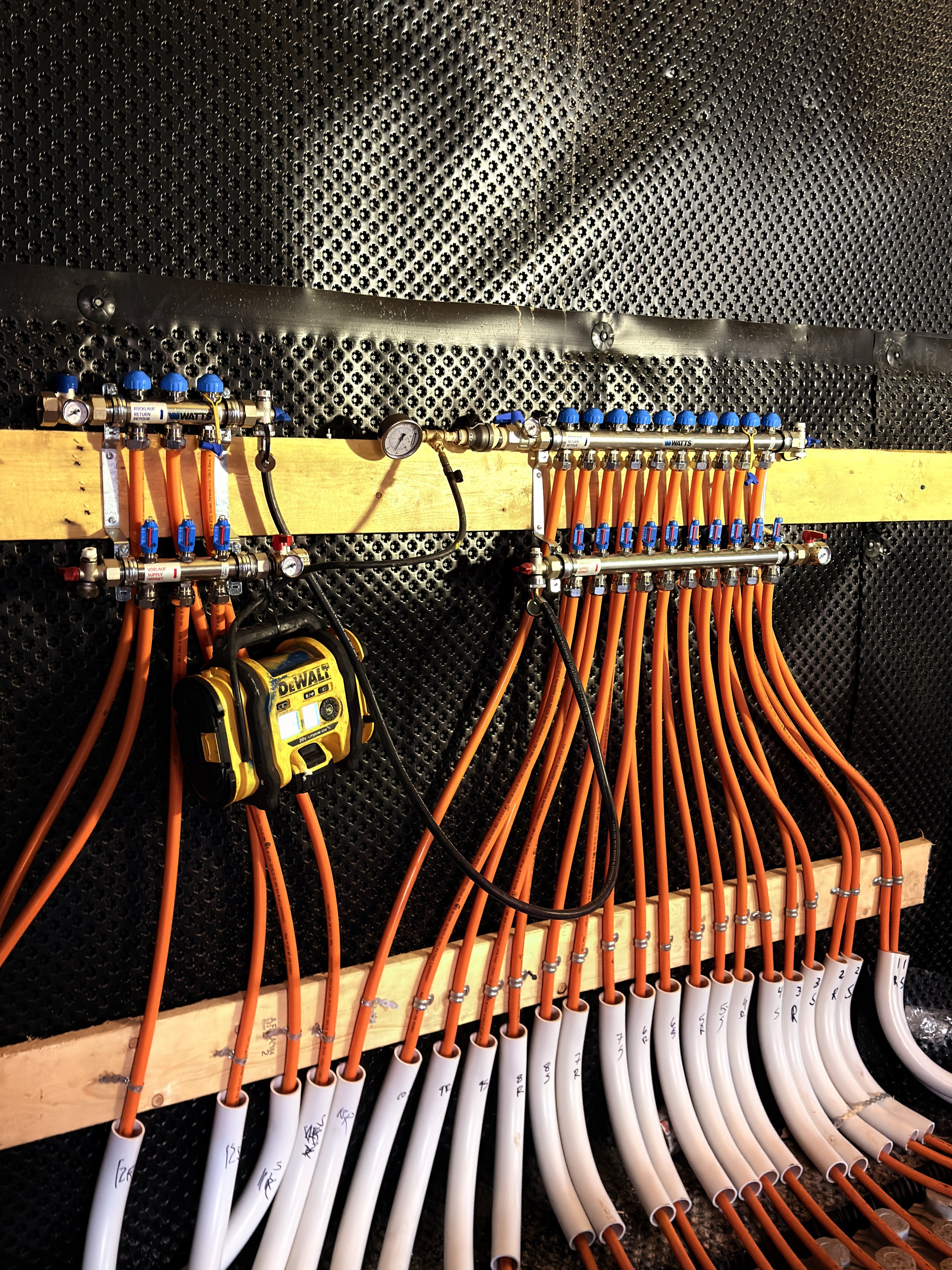 Electrical or plumbing piping system with colored pipes, gauges, and valves mounted on a black perforated wall, with a yellow measuring device attached.