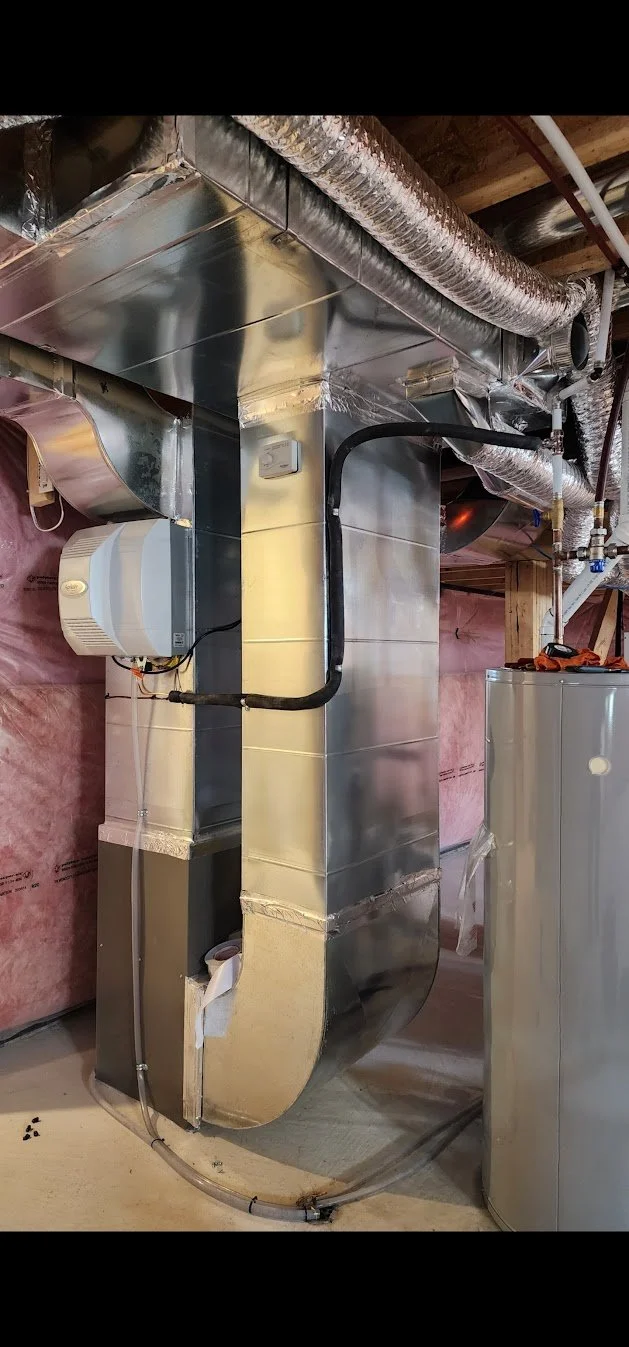 Metal HVAC duct system with flexible insulated ducting, mounted on a basement wall with pink insulation, next to a water heater.
