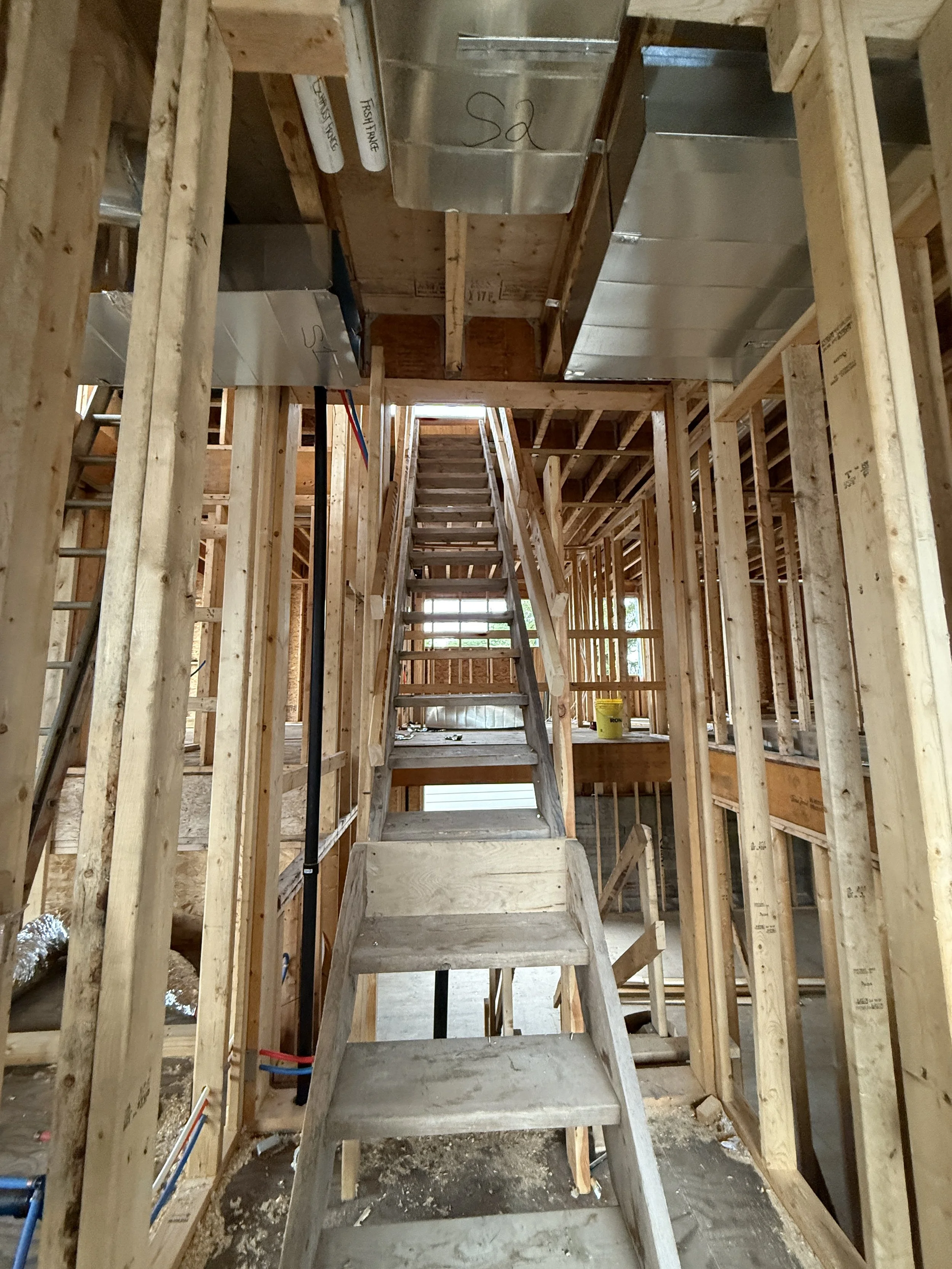 Interior view of a house under construction showing wooden framing, a staircase leading to the upper level, and construction materials.