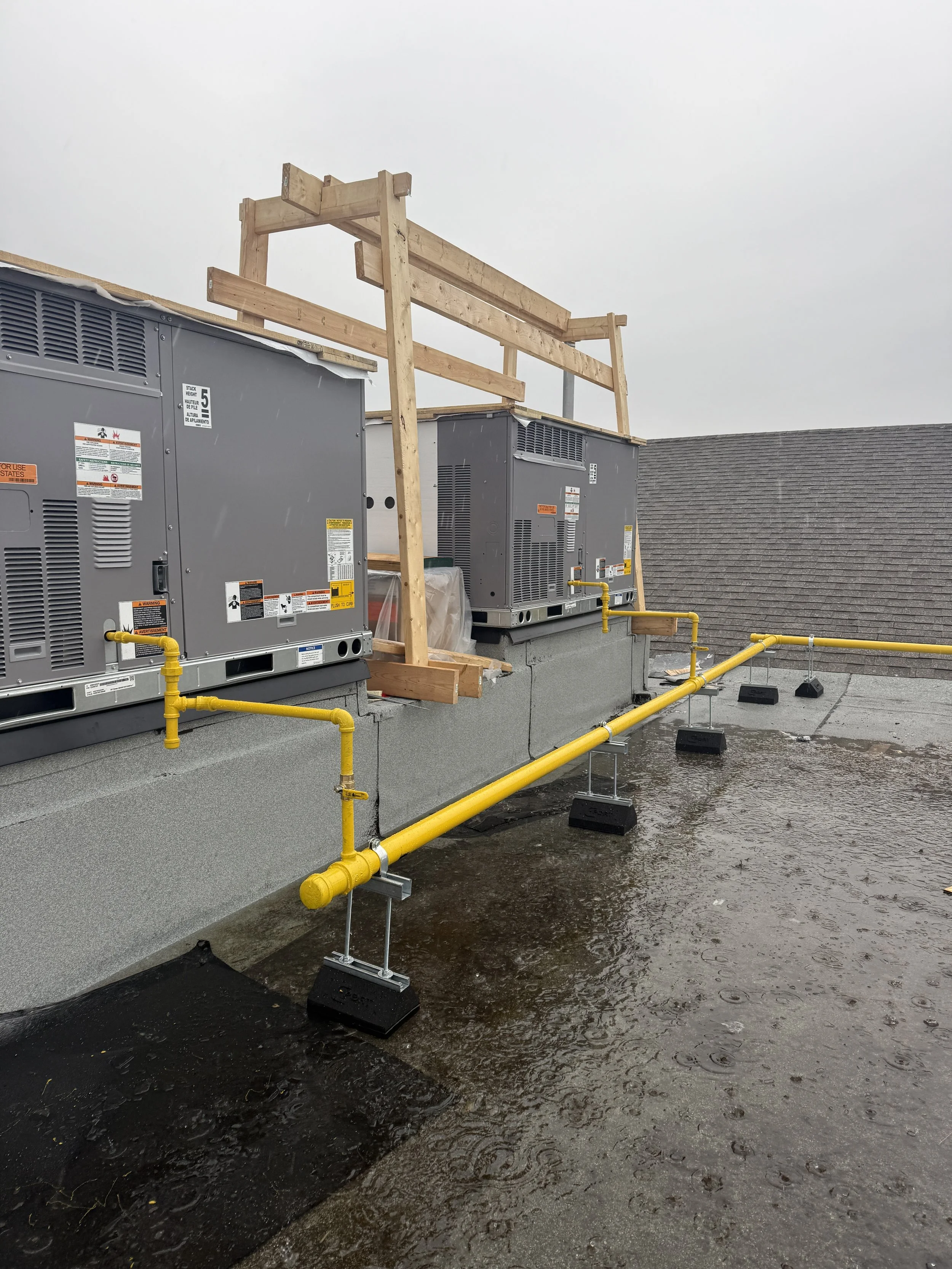 Rooftop HVAC units with a temporary wood guardrail and a yellow gas line, on a rainy day.