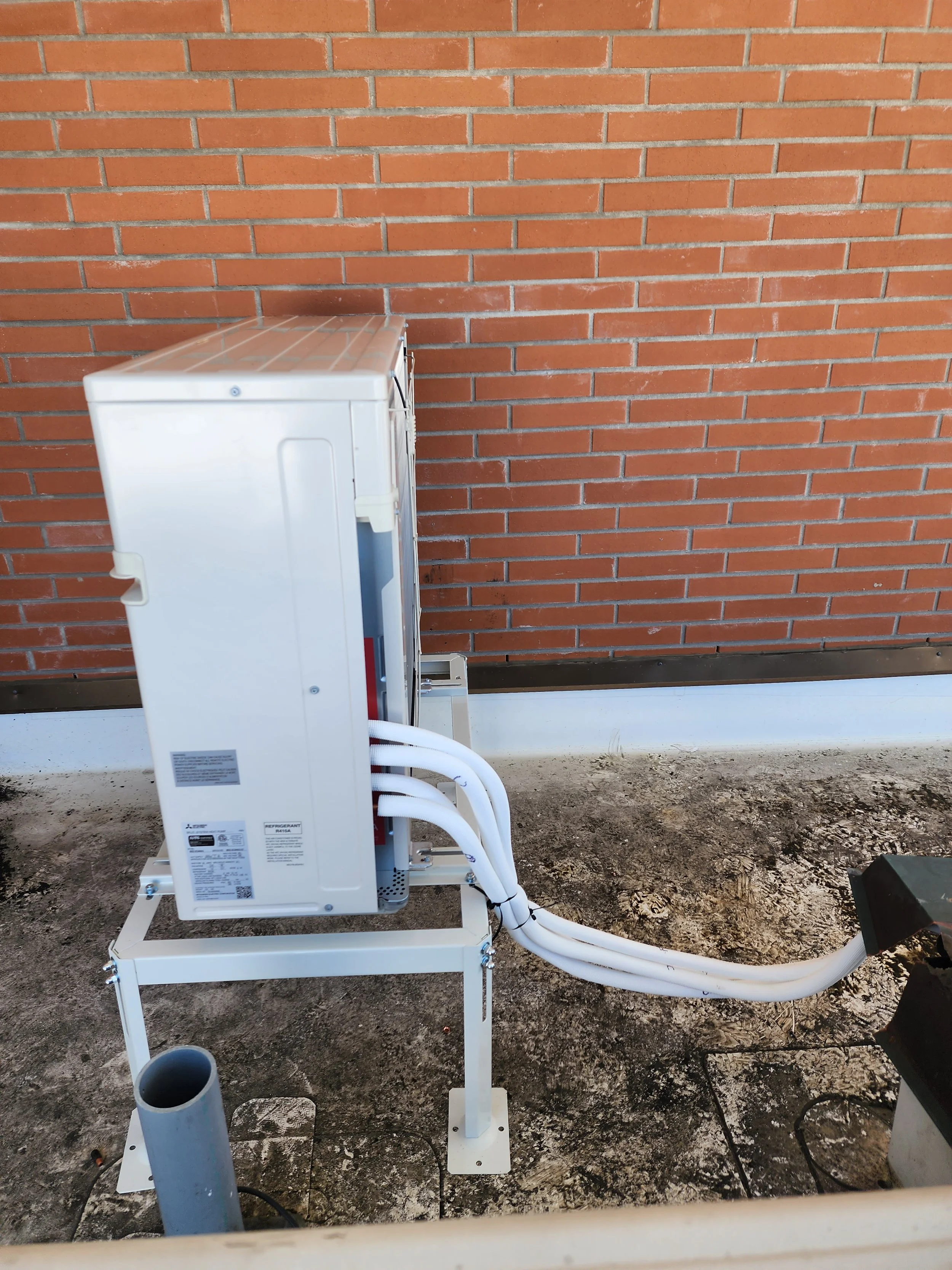 An outdoor HVAC unit installed on a metal stand against a brick wall, with insulated pipes extending from it.