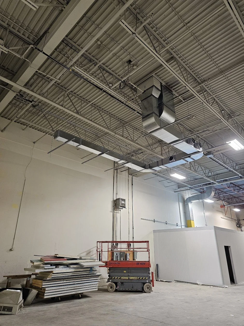 Construction site with a high ceiling, exposed ductwork and ceilings, a small red lift, and a white temporary wall or booth.