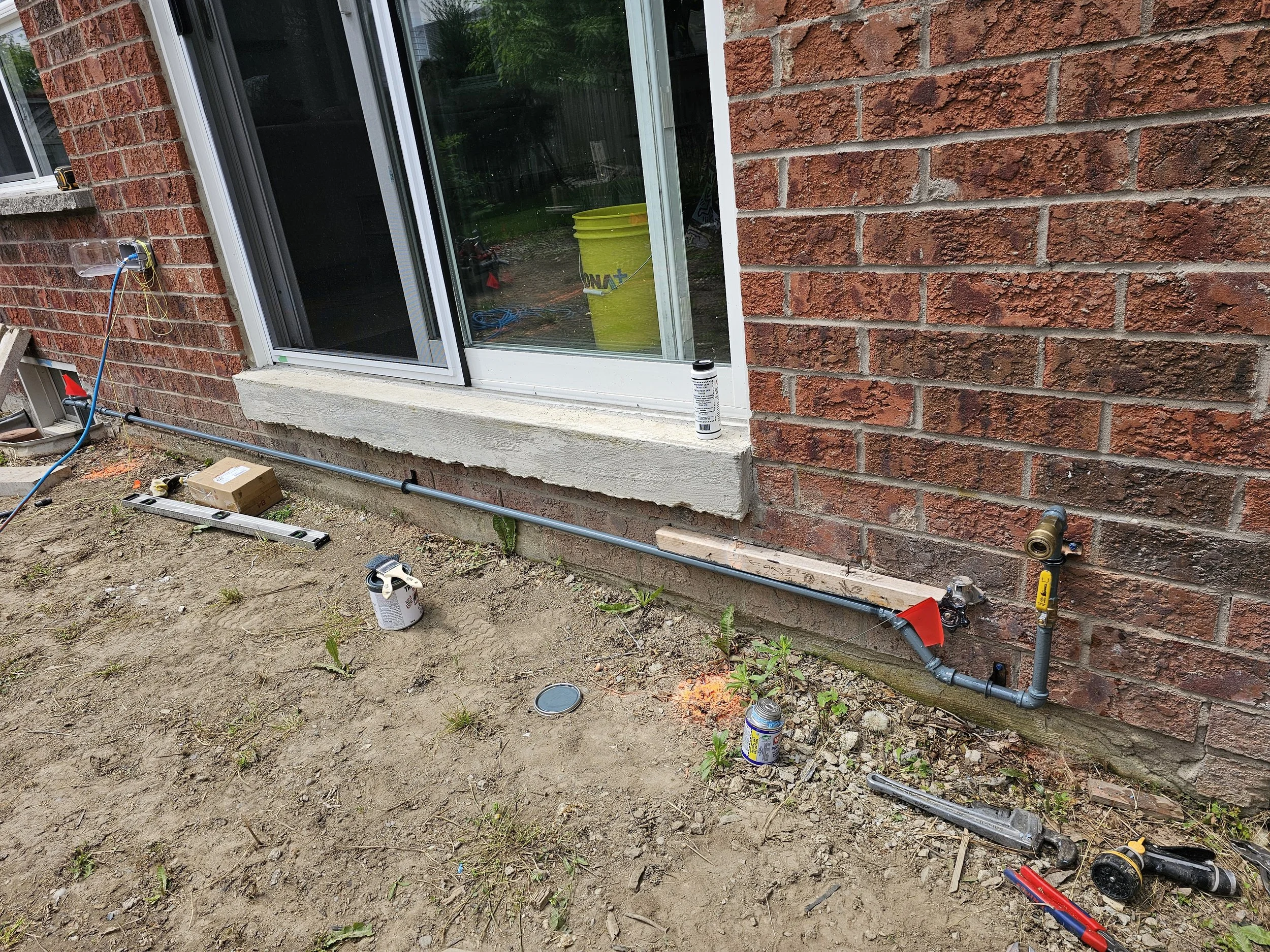 Installation of an outdoor water supply line on the side of a brick house, with tools and pipes laid out on the ground.