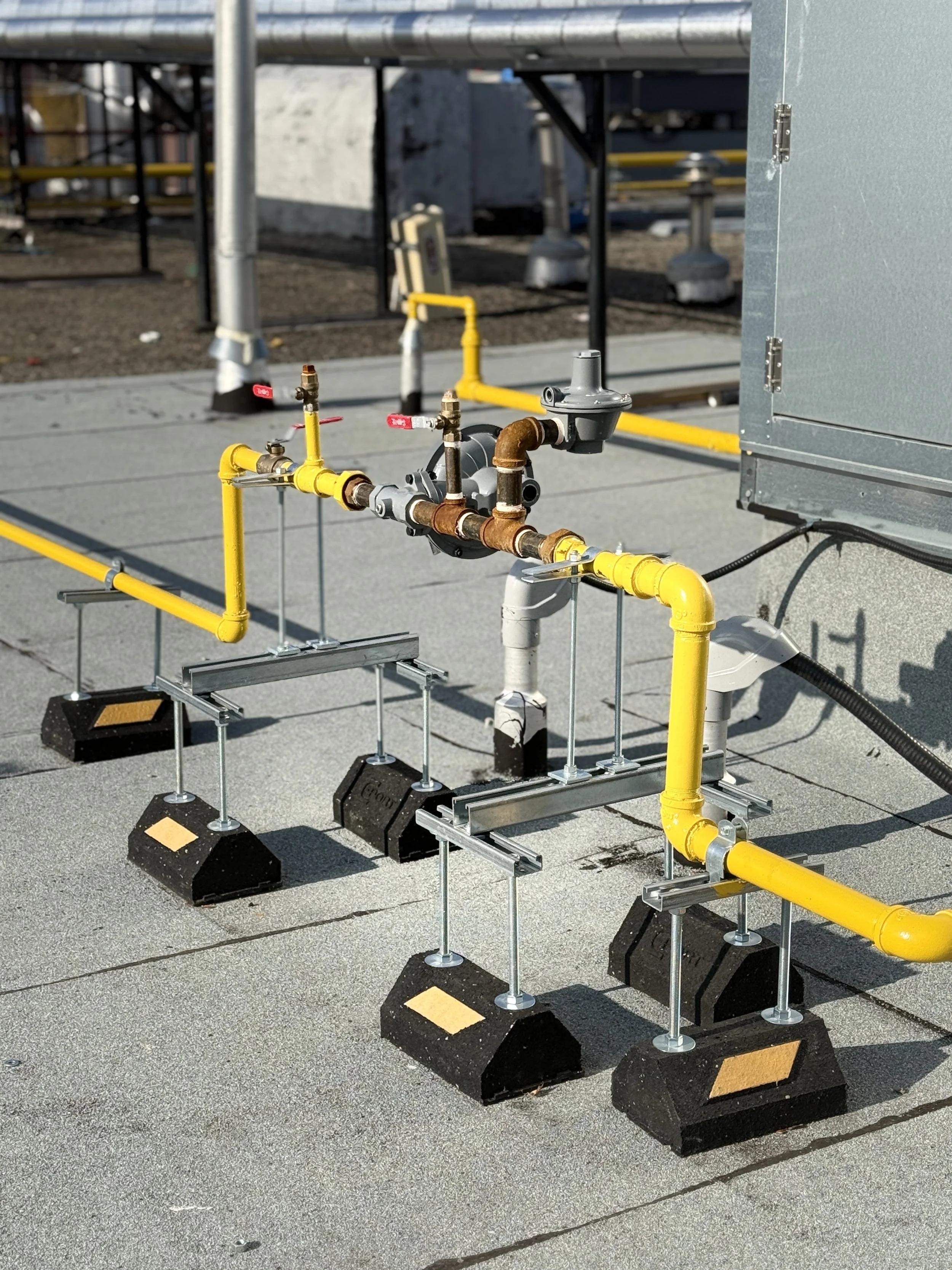Set of gas pipes and valves on a rooftop, secured with metal supports and concrete blocks, with electrical equipment and piping in the background.