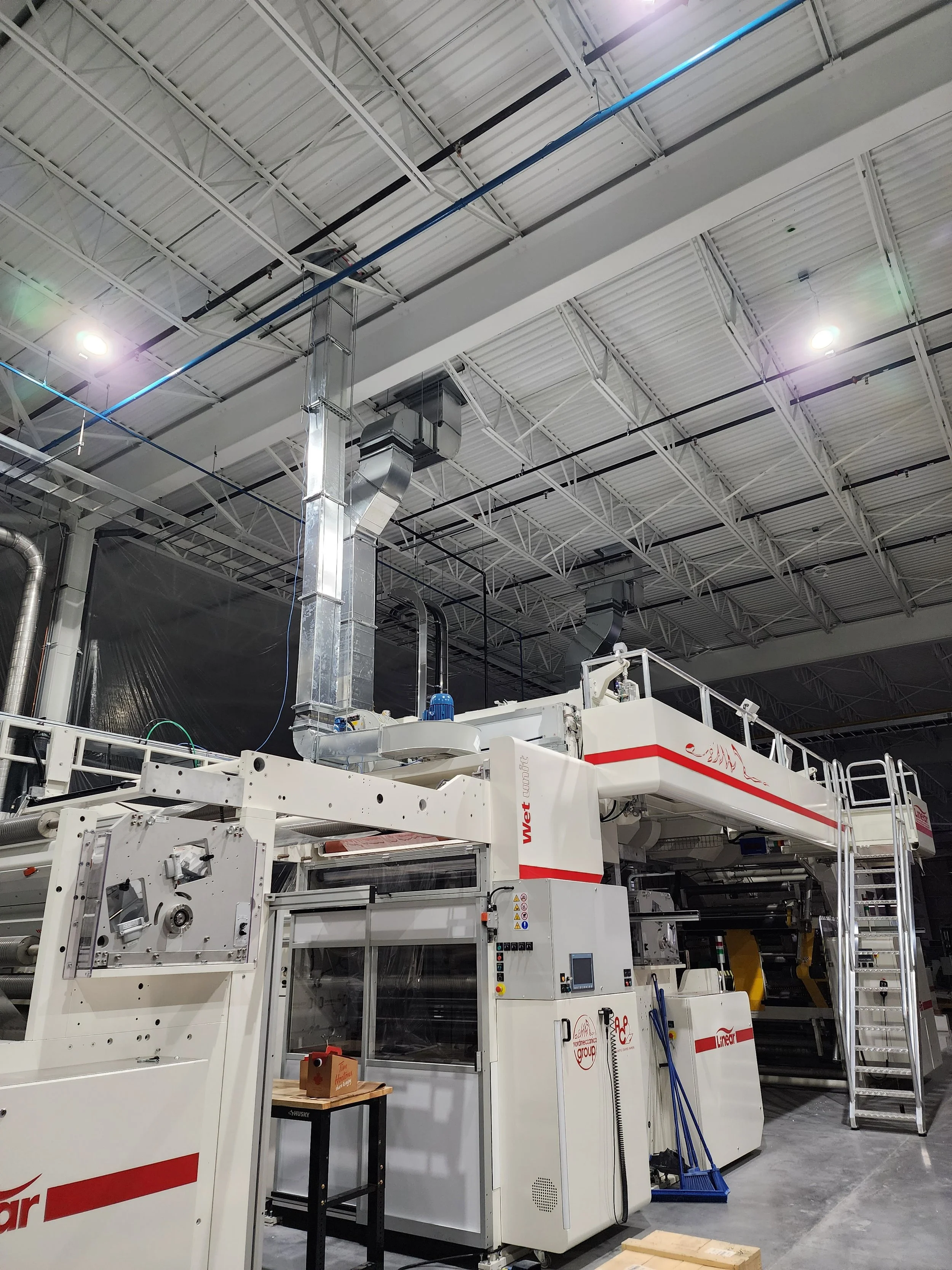 Large industrial machine with metal stairs and control panel, located in a warehouse with high ceiling and metal beams.