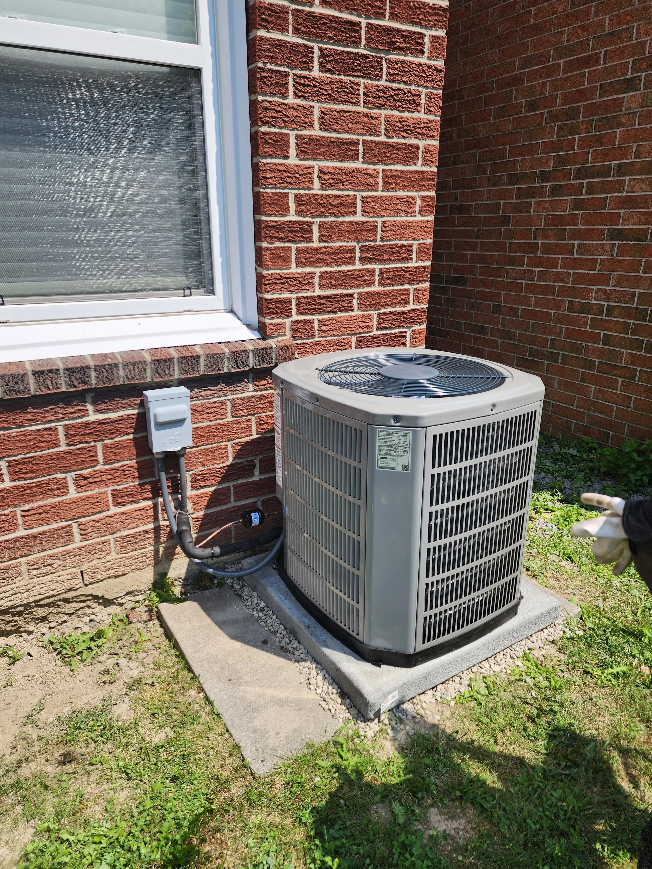 Outdoor air conditioning unit next to a brick house wall, installed on a concrete pad, with a small garden area and a window with closed blinds.