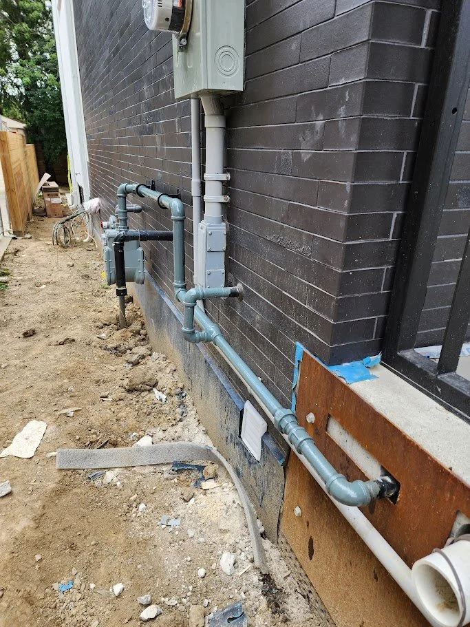Outside brick wall of a house with plumbing pipes and electrical box installed on the foundation, and construction materials scattered on the ground.