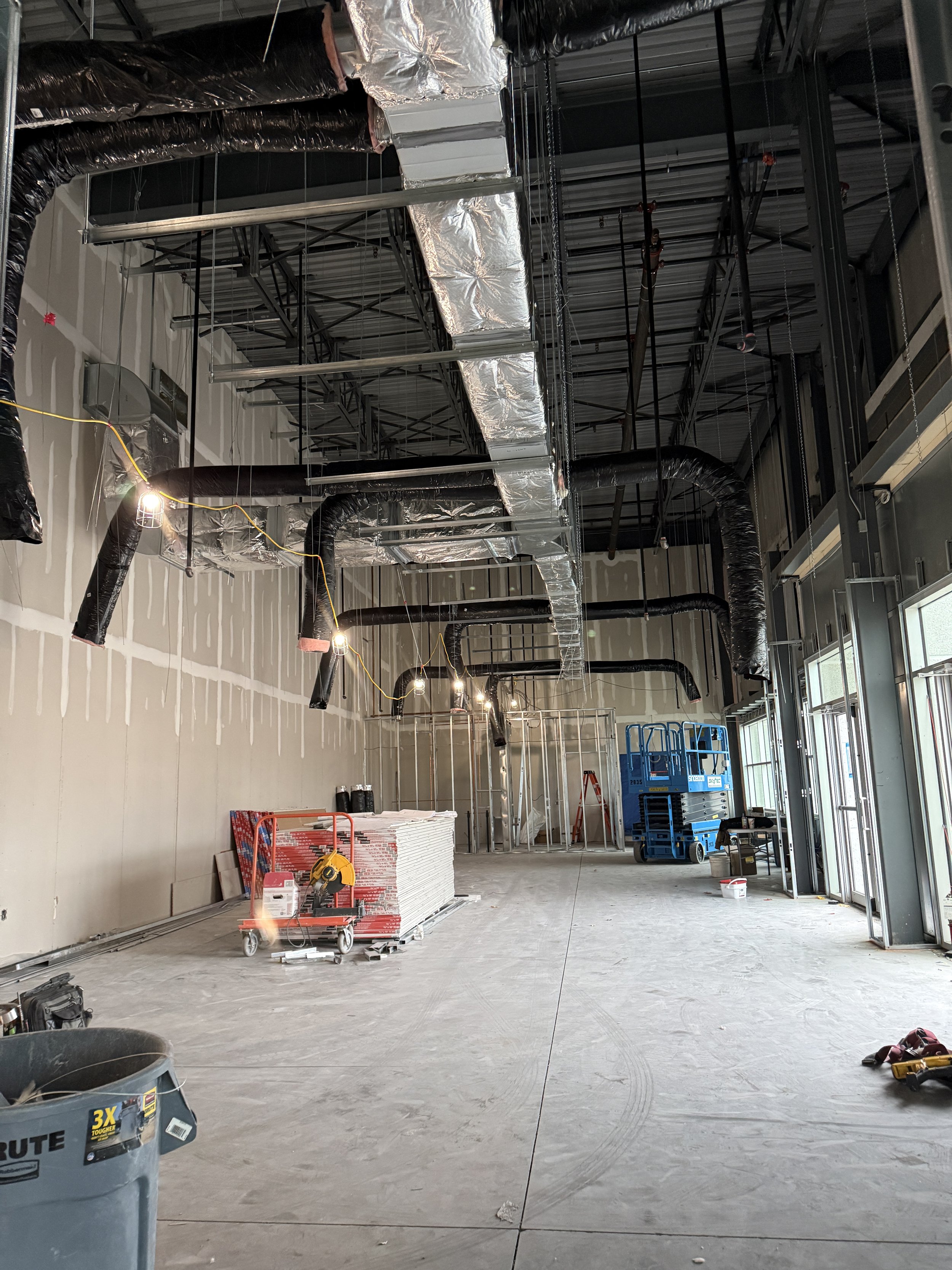 Interior view of a commercial building under construction with exposed ductwork, electrical wiring, and framing. Construction tools and materials are scattered on the unfinished concrete floor, with a blue scissor lift along the right wall.