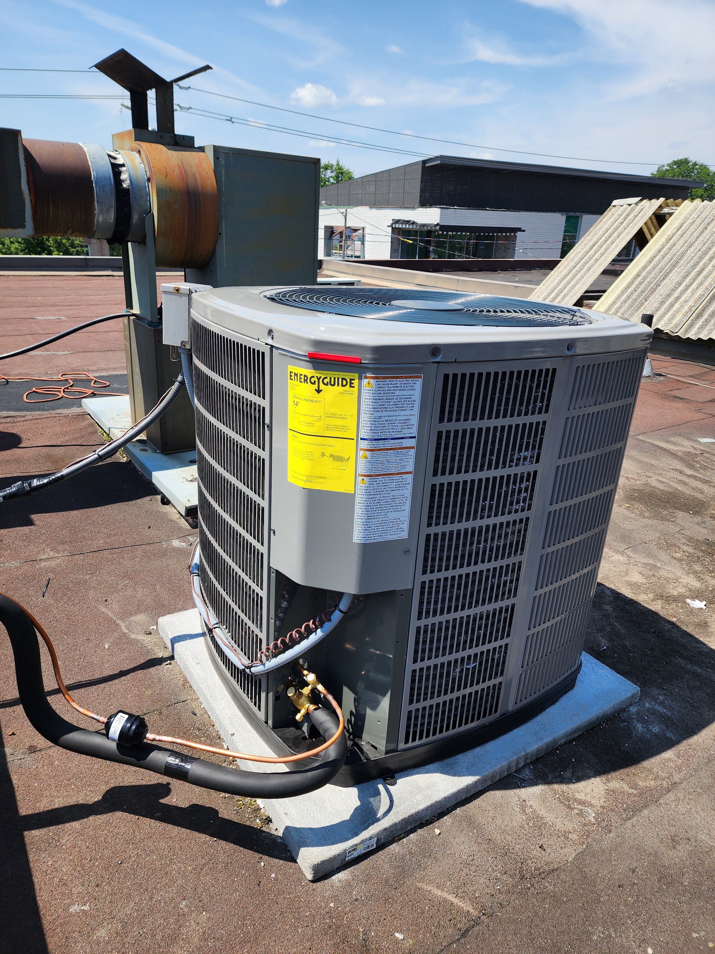 HVAC unit on a rooftop with a yellow Energy Guide sticker, copper pipes, and a vent pipe in the background.