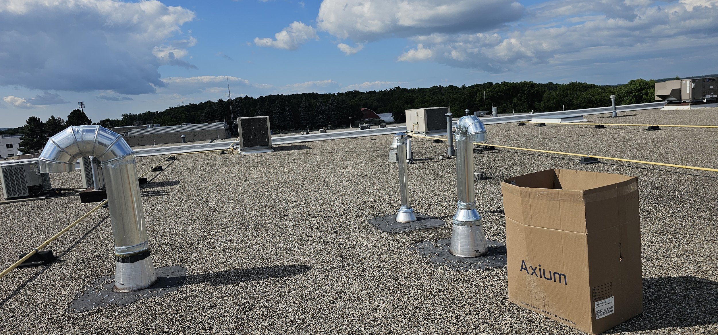 Rooftop with HVAC units, metallic air vents, yellow security lines, and a cardboard box with the label 'Axiom' under a partly cloudy sky.