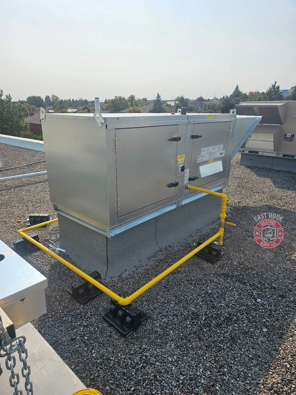 Rooftop HVAC unit installed on a gravel surface, surrounded by yellow piping and black mounting blocks, with trees and houses in the background.