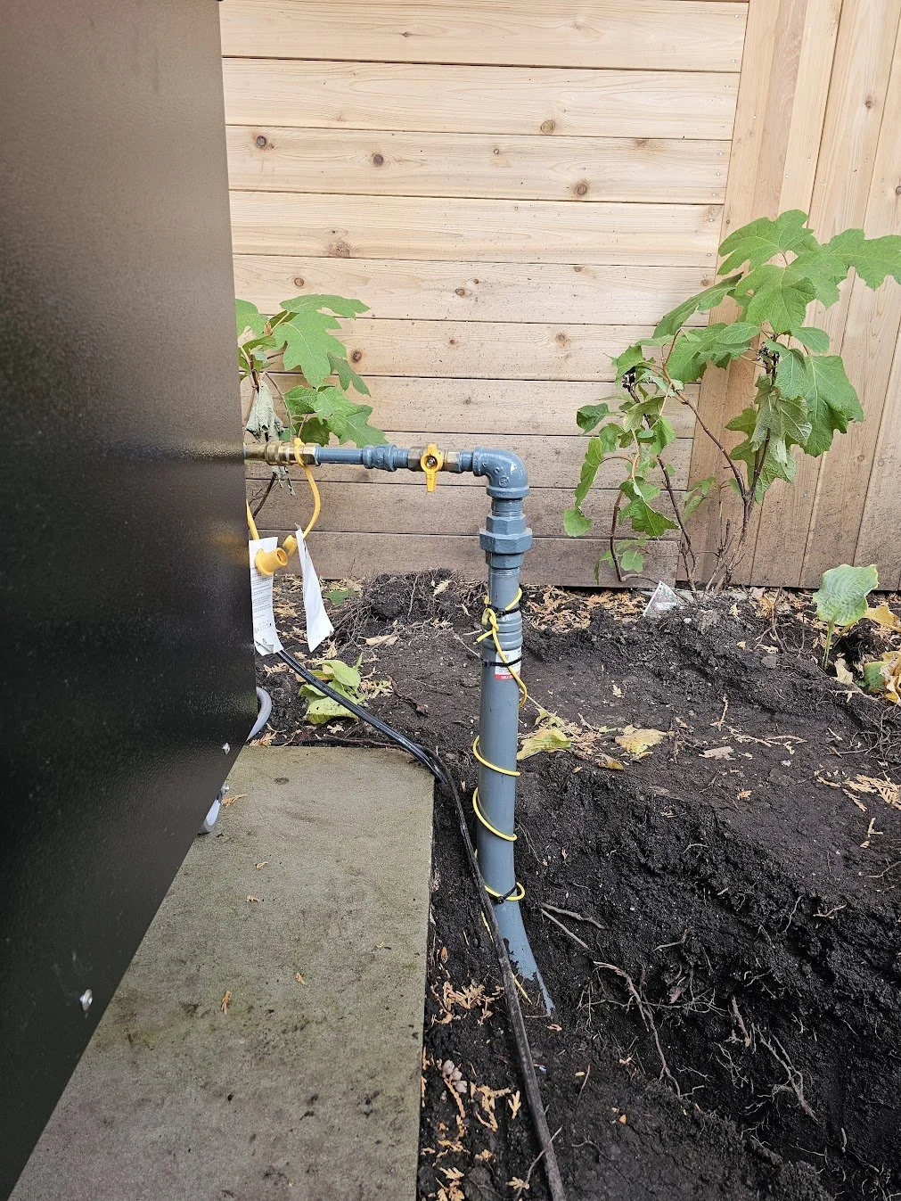 Outdoor water meter with piping and electronics, situated in a garden with soil and green plants, against a wooden fence and wall.