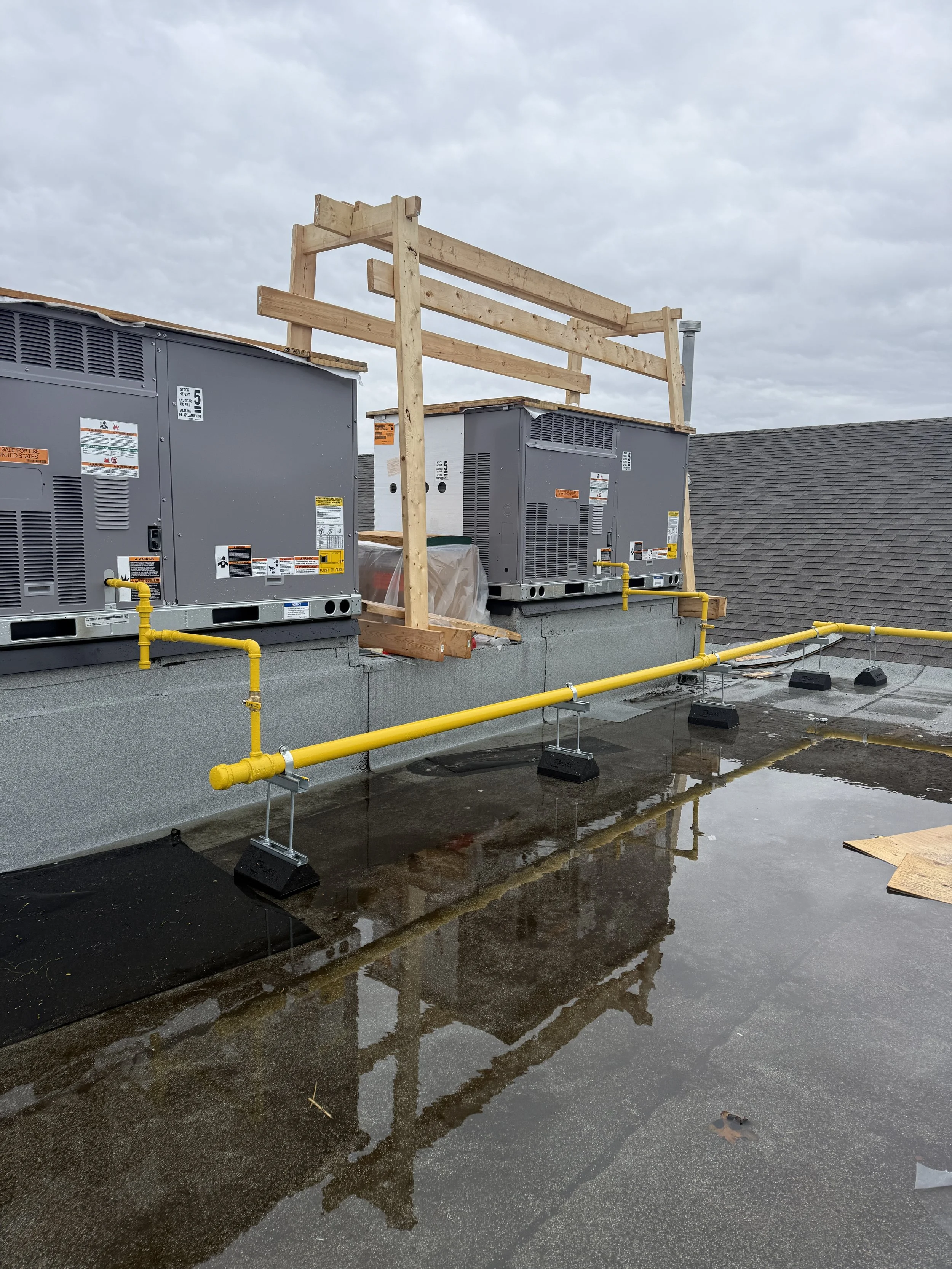 Rooftop with construction work including air conditioning units, yellow piping, and wooden framing, with puddles of water and a cloudy sky.