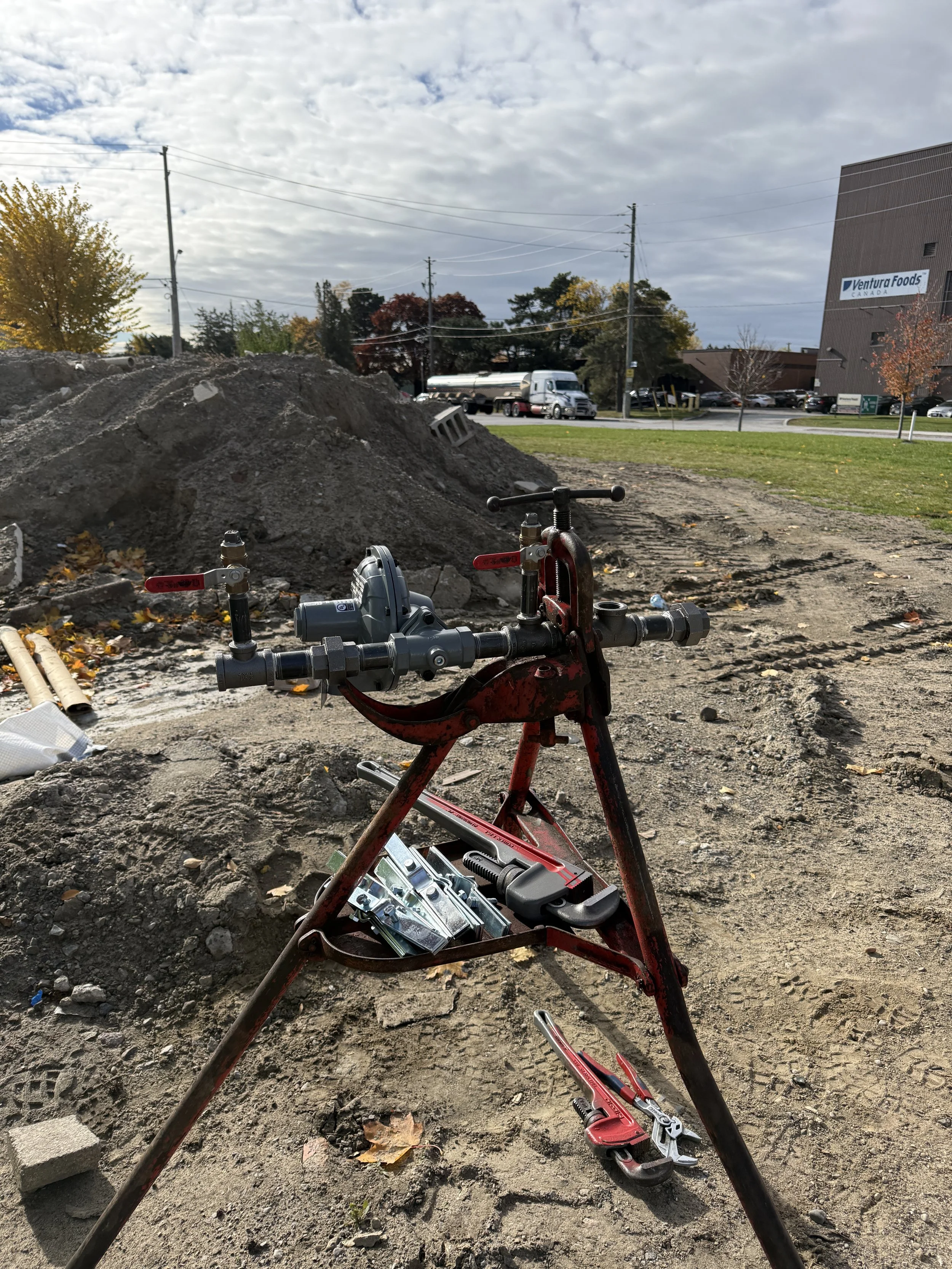 Construction site with a hydraulic pipe press and tools on disturbed dirt, background features a large pile of soil, utility poles, trees with fall foliage, and a building labeled Ventura Foods.