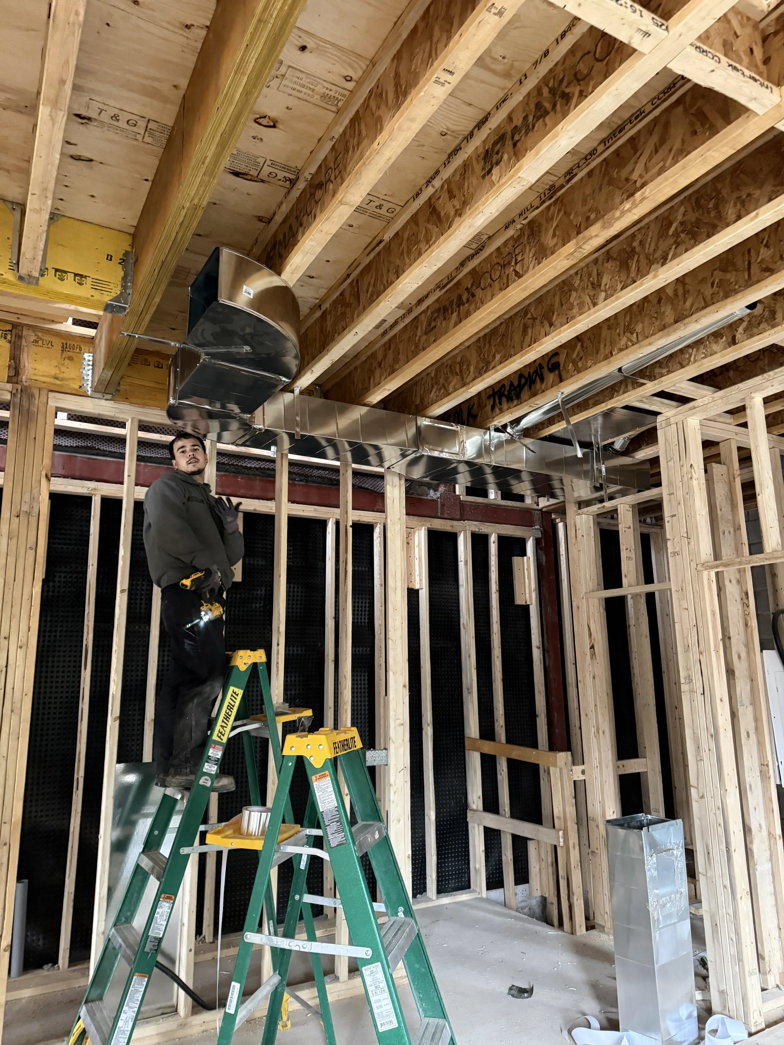 A construction worker standing on a green ladder inside a building under construction, installing or working near ductwork on the ceiling, with wooden framing and partially installed roof.