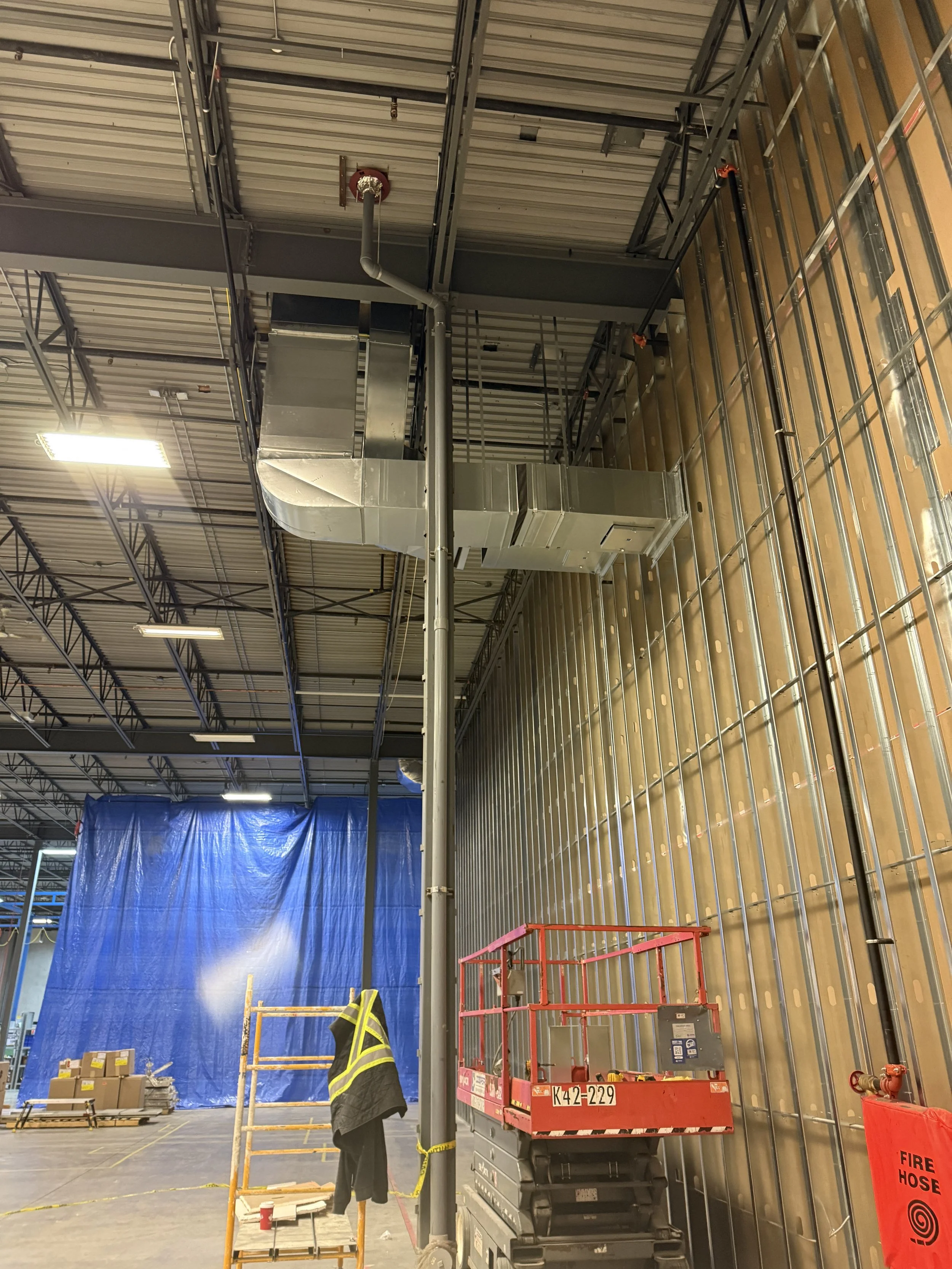 Construction inside an industrial building with ductwork, a scissor lift, a high-visibility vest hanging on a scaffold, and a fire hose cabinet.