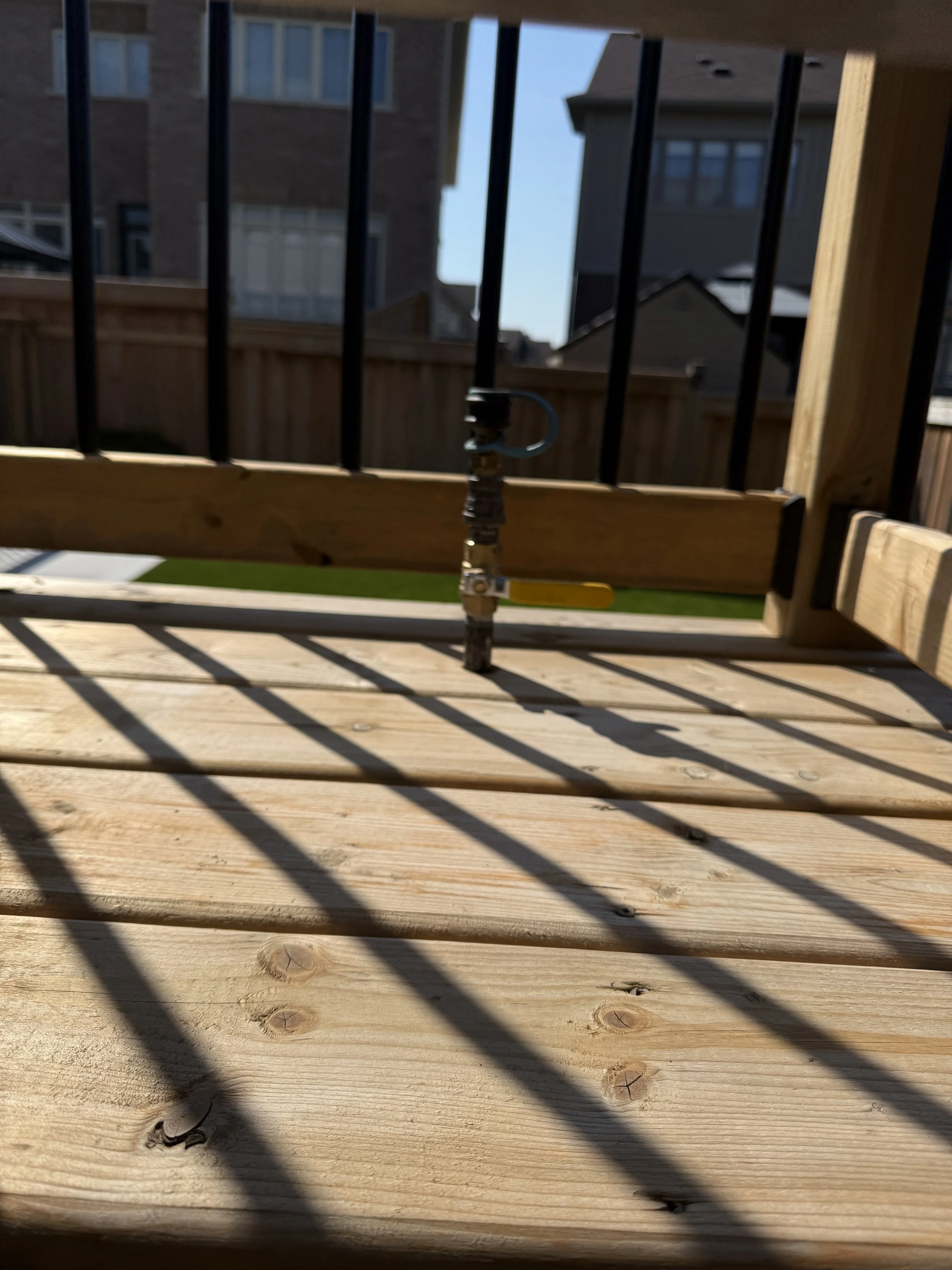 The photo shows a close-up view of a wooden deck with shadows cast by a railing. There is a bolt or clamp in the center attached to a yellow handle, possibly a part of the deck construction or a tool, with a background of neighboring houses and a blue sky.